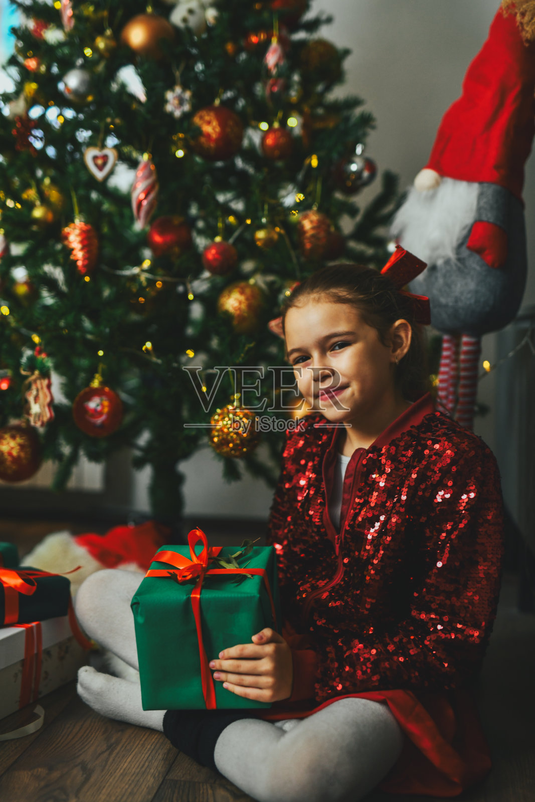 穿着红色闪亮服装的美丽女孩坐在圣诞树前，腿上放着包好的礼物。照片摄影图片