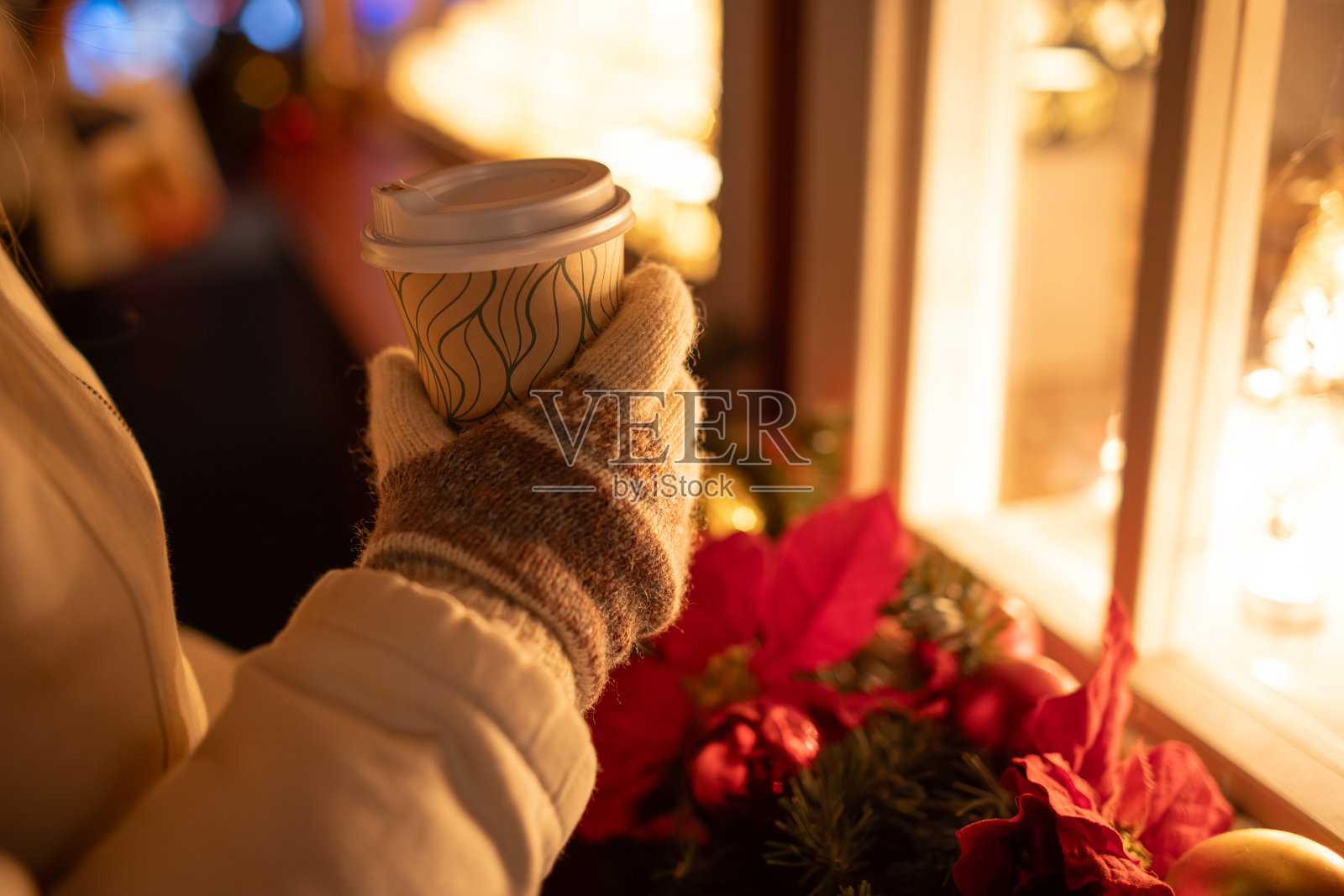 在欧洲的圣诞市场上，一只戴着冬季手套的女性手握着一个普通的外带塑料咖啡杯。特写镜头，浅景深，夜晚，装饰灯光。照片摄影图片