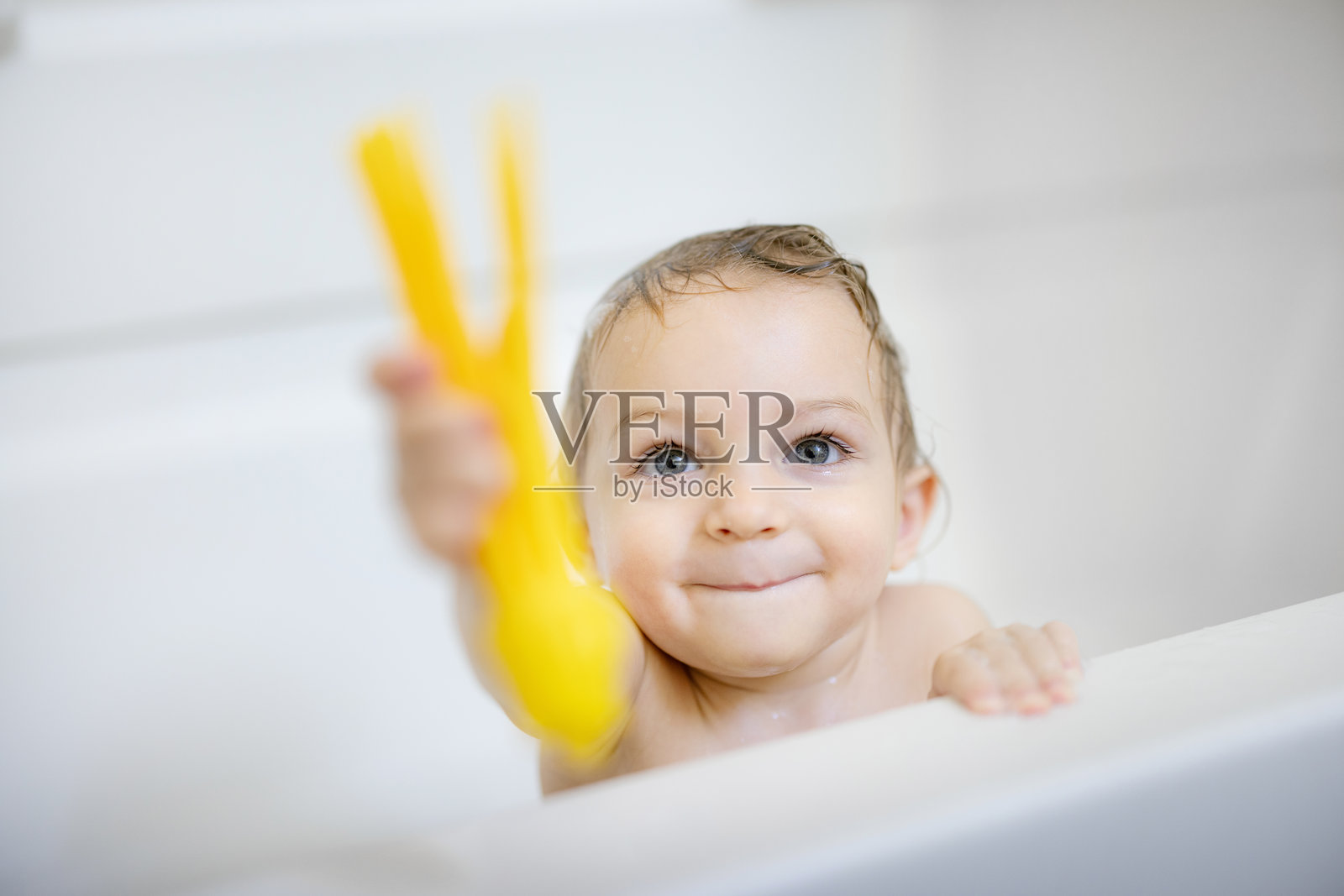 可爱的宝宝在浴缸里玩耍，手里拿着一个黄色玩具，明亮的光线照耀着。照片摄影图片
