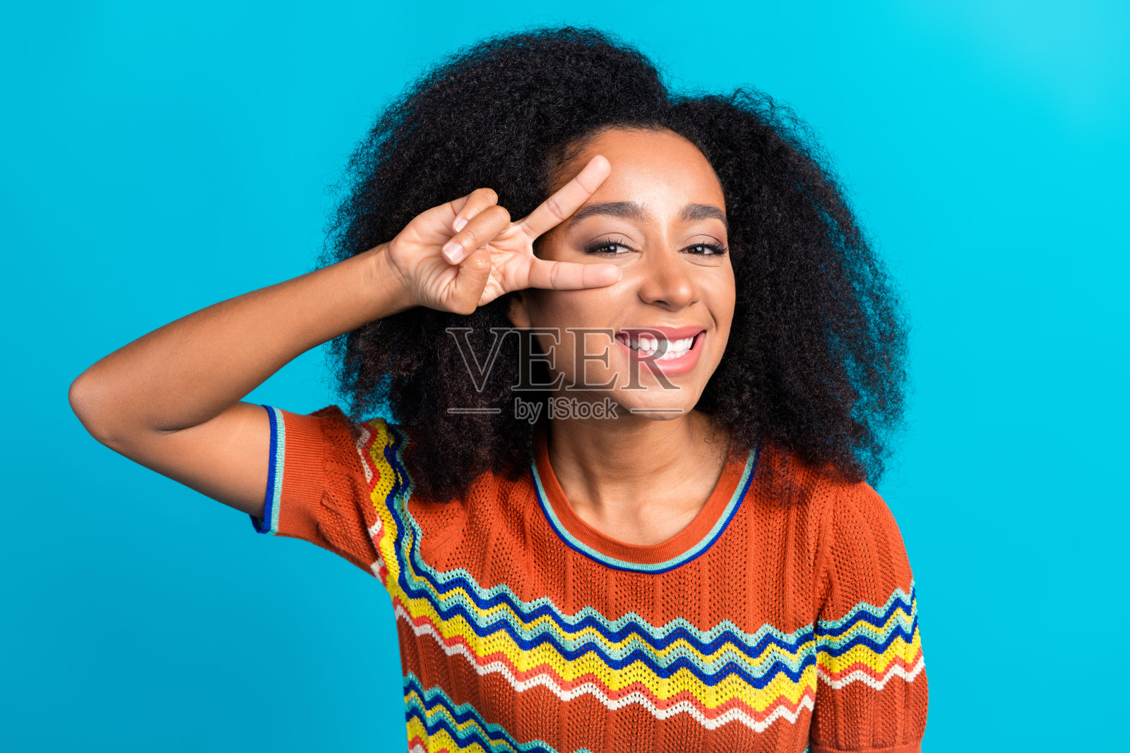微笑的年轻女性，蓬松的非洲卷发，背景为鲜艳的蓝色，做出和平手势照片摄影图片