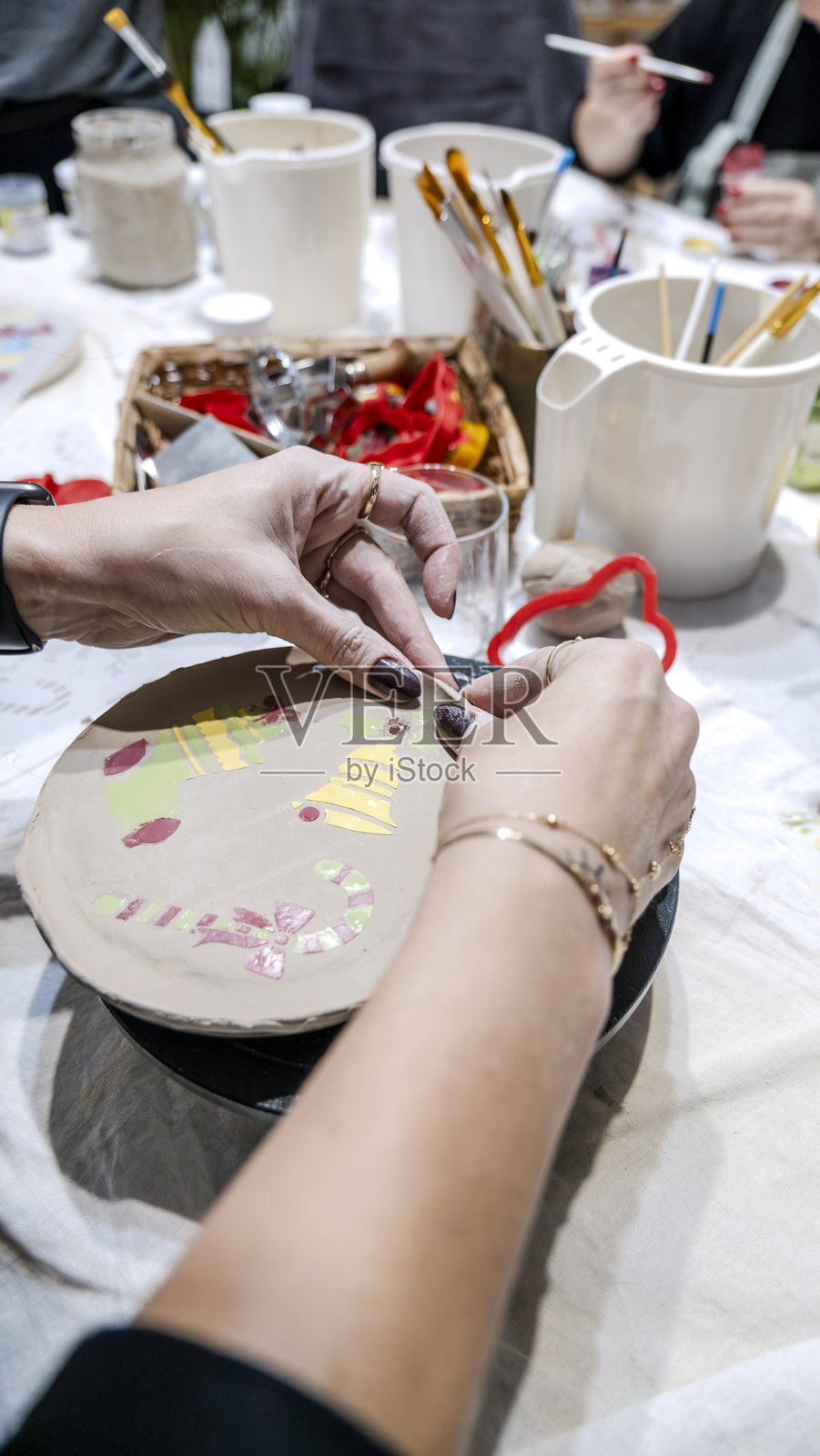 一位中年女性艺术家在艺术工作室的桌子上用雕刻工具塑形粘土，背景是装饰着艺术画作的环境。照片摄影图片