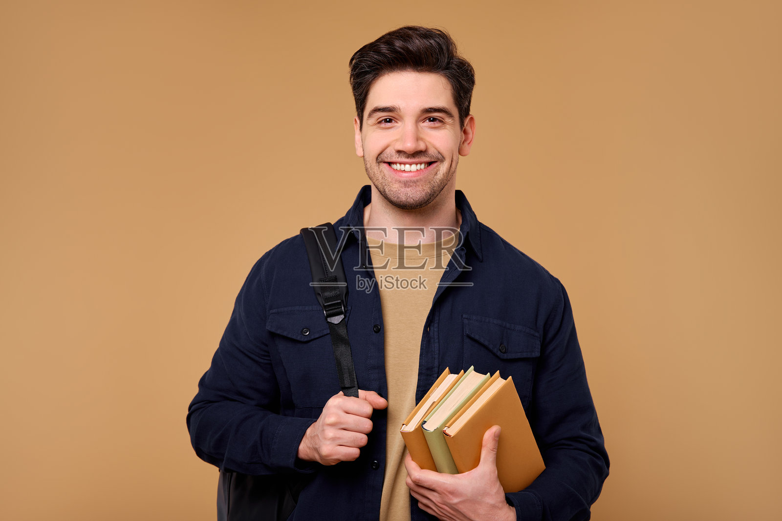 一个快乐时尚的男人背着书包，微笑着站在米色背景前照片摄影图片