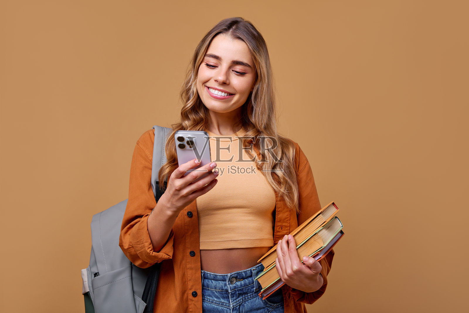 一个快乐的学生手持书籍和智能手机，站在温馨舒适的背景前。照片摄影图片