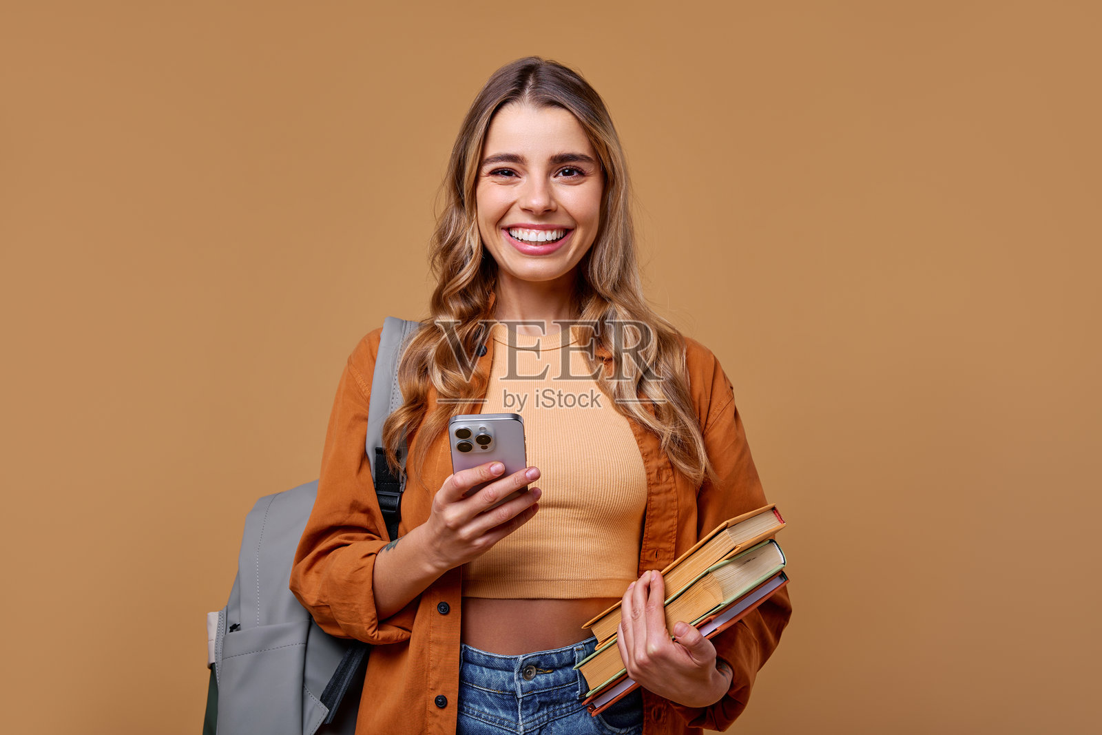 一个快乐的学生手持书籍和智能手机，站在温馨舒适的背景前。照片摄影图片