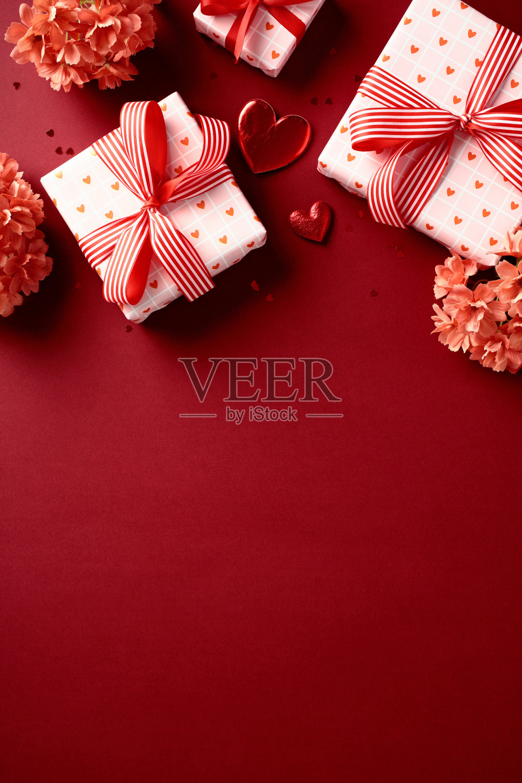 情人节礼品盒，配有心形图案的纸和红色丝带，周围环绕着红色花朵和心形装饰，背景为红色竖条纹。照片摄影图片