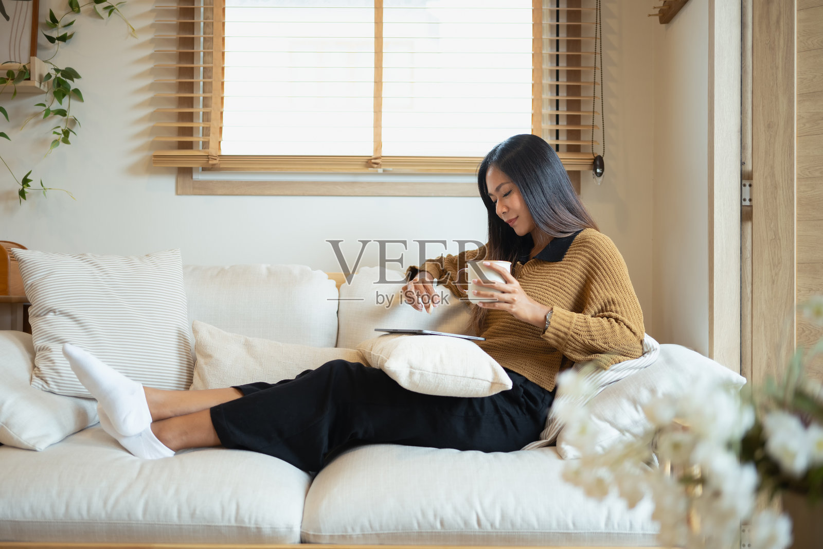 美丽的年轻女性坐在柔软的沙发上，手里拿着一个杯子，正在使用平板电脑。照片摄影图片