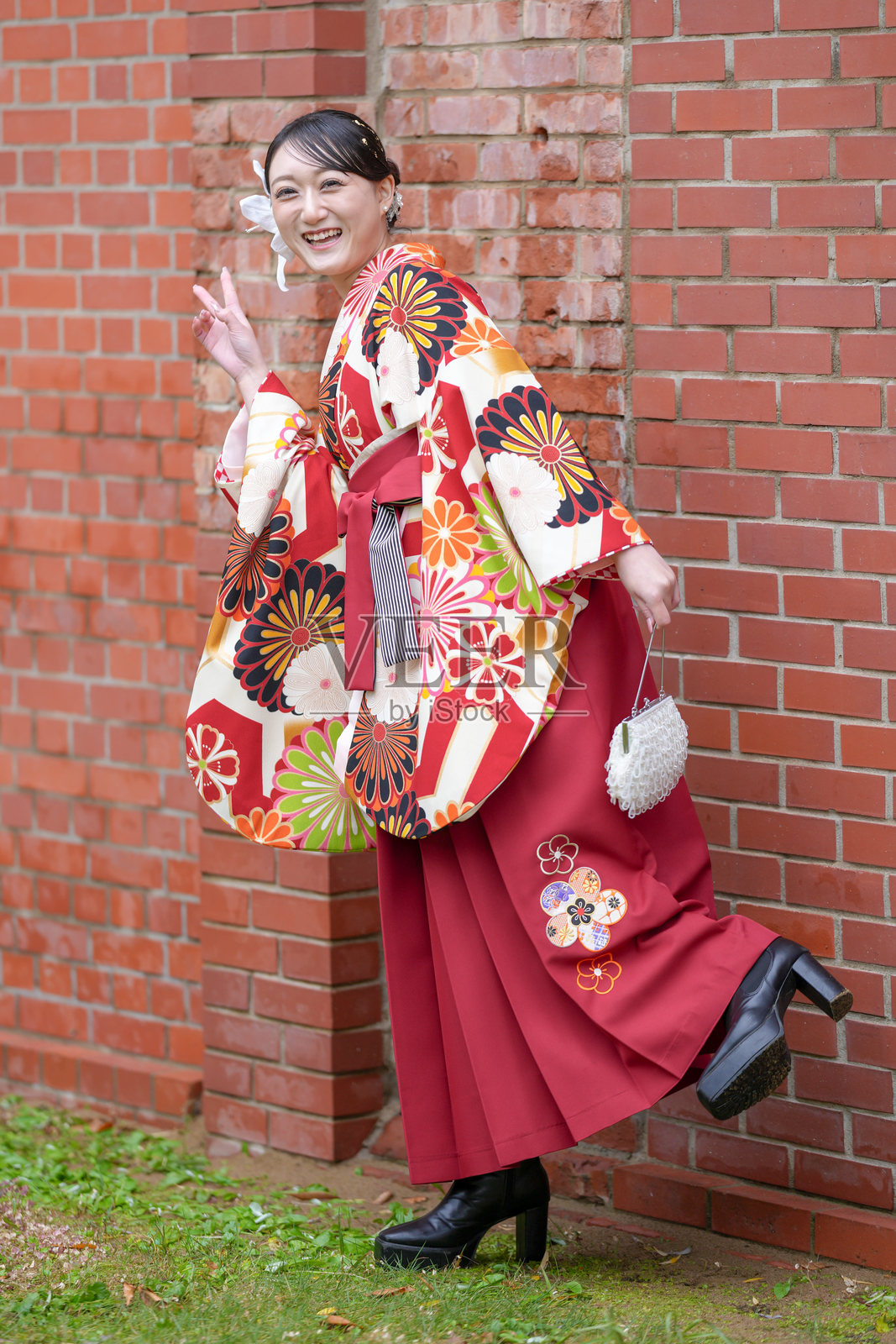 一位20多岁的日本女性，穿着红色袴，作为日本大学毕业生的标志，站在一座文化砖砌建筑前摆姿势。照片摄影图片