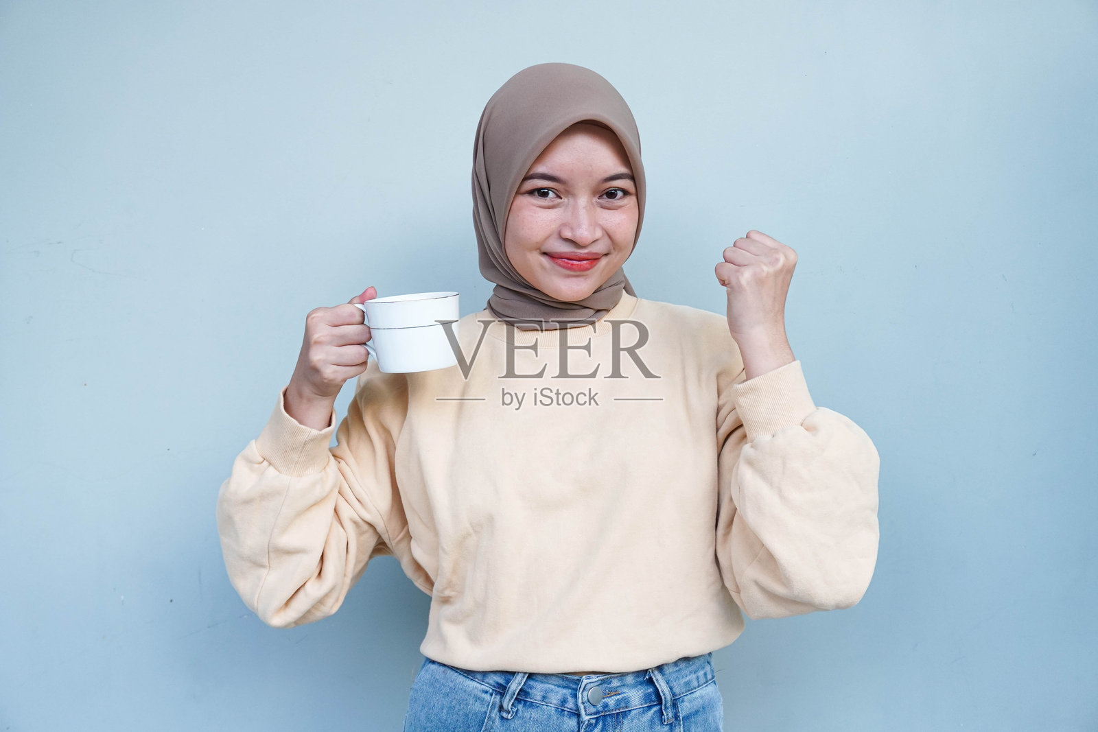 快乐美丽的亚洲穆斯林女性穿着奶油色毛衣，手握咖啡杯，庆祝好运，背景为白色。照片摄影图片