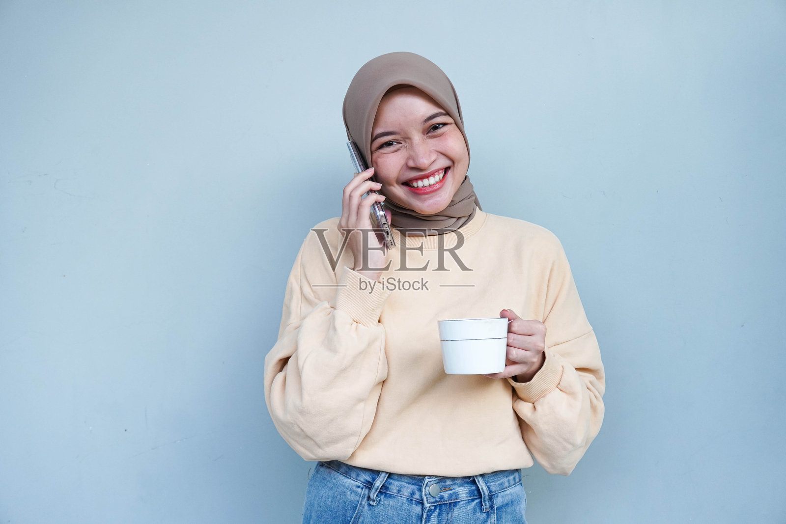 微笑的美丽亚洲穆斯林女性，穿着奶油色毛衣和头巾，侧身看着一旁，正在用手机通话，背景为白色。照片摄影图片