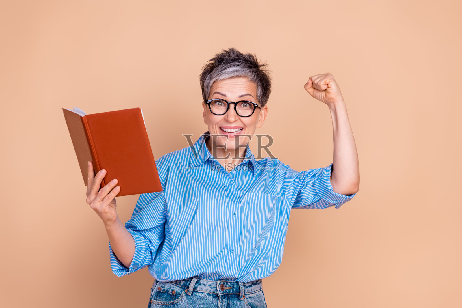 自信的成熟的女性，短灰发，举起拳头欢呼，手中拿着一本书，背景为米色。照片摄影图片