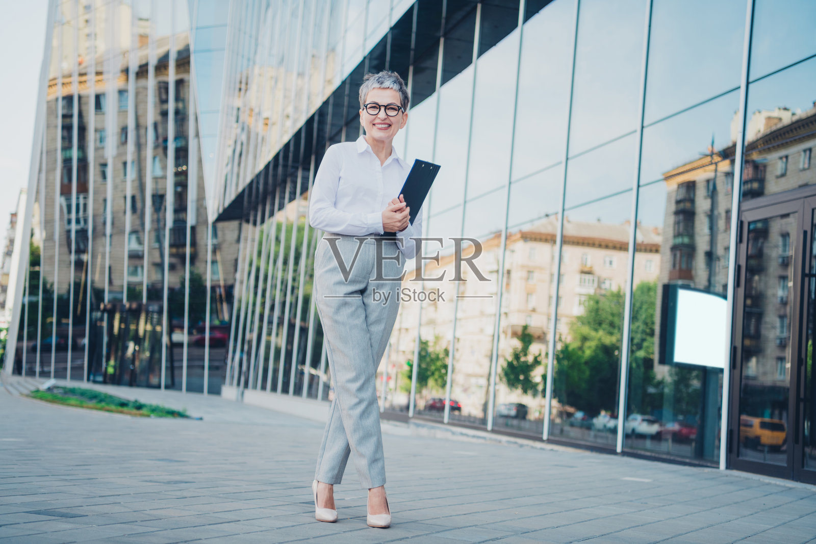自信的成熟的女商人在现代建筑和反射窗户的城市景观中穿着正式服装站立。照片摄影图片