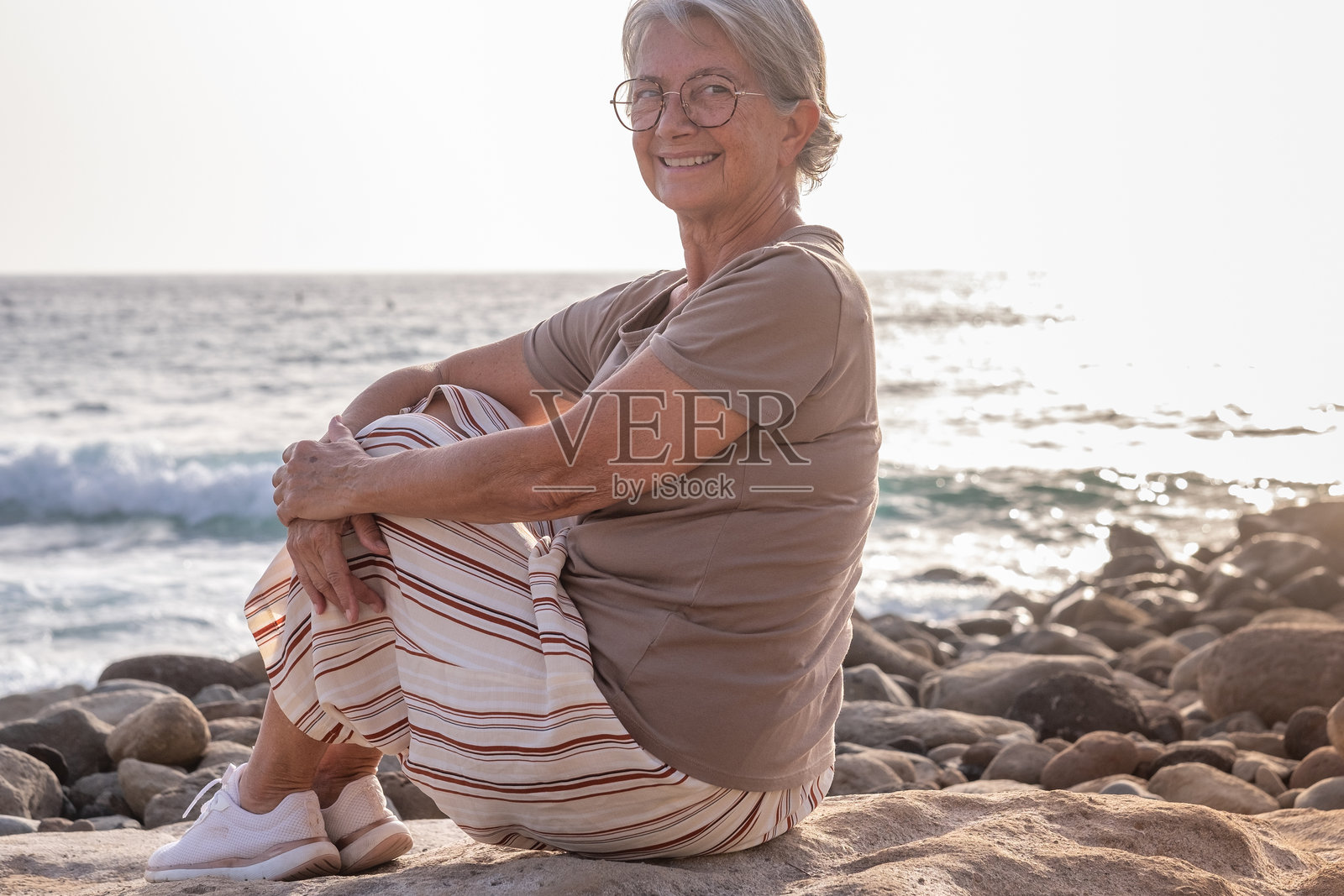 迷人的放松的老年女性坐在海滩上，夕阳下微笑着看向镜头 - 老年女士享受假期和自由照片摄影图片