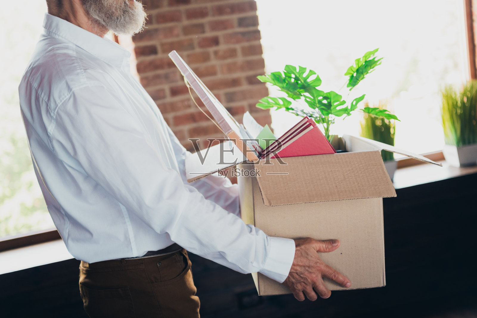 裁剪的照片，展示了一位和蔼的老年人穿着白色衬衫和正式服装，手里提着纸箱，在明亮的办公室中与同事们合作，展现出成功的气息。照片摄影图片