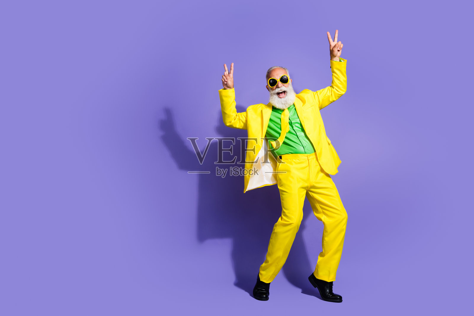全身肖像的搞笑奢华老年男子，做出V字手势，身穿黄色西装，背景为紫色。照片摄影图片