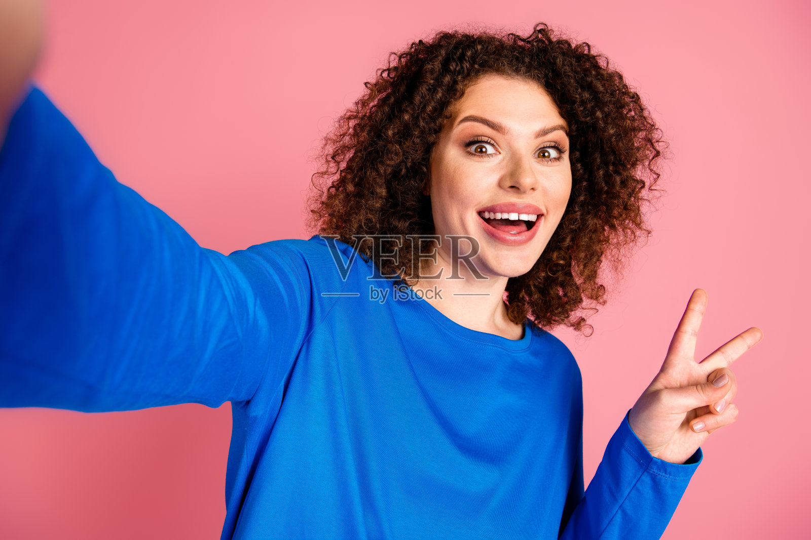 穿着蓝色卫衣的年轻女性在粉色背景前摆姿势，做出和平手势，面带灿烂笑容。照片摄影图片