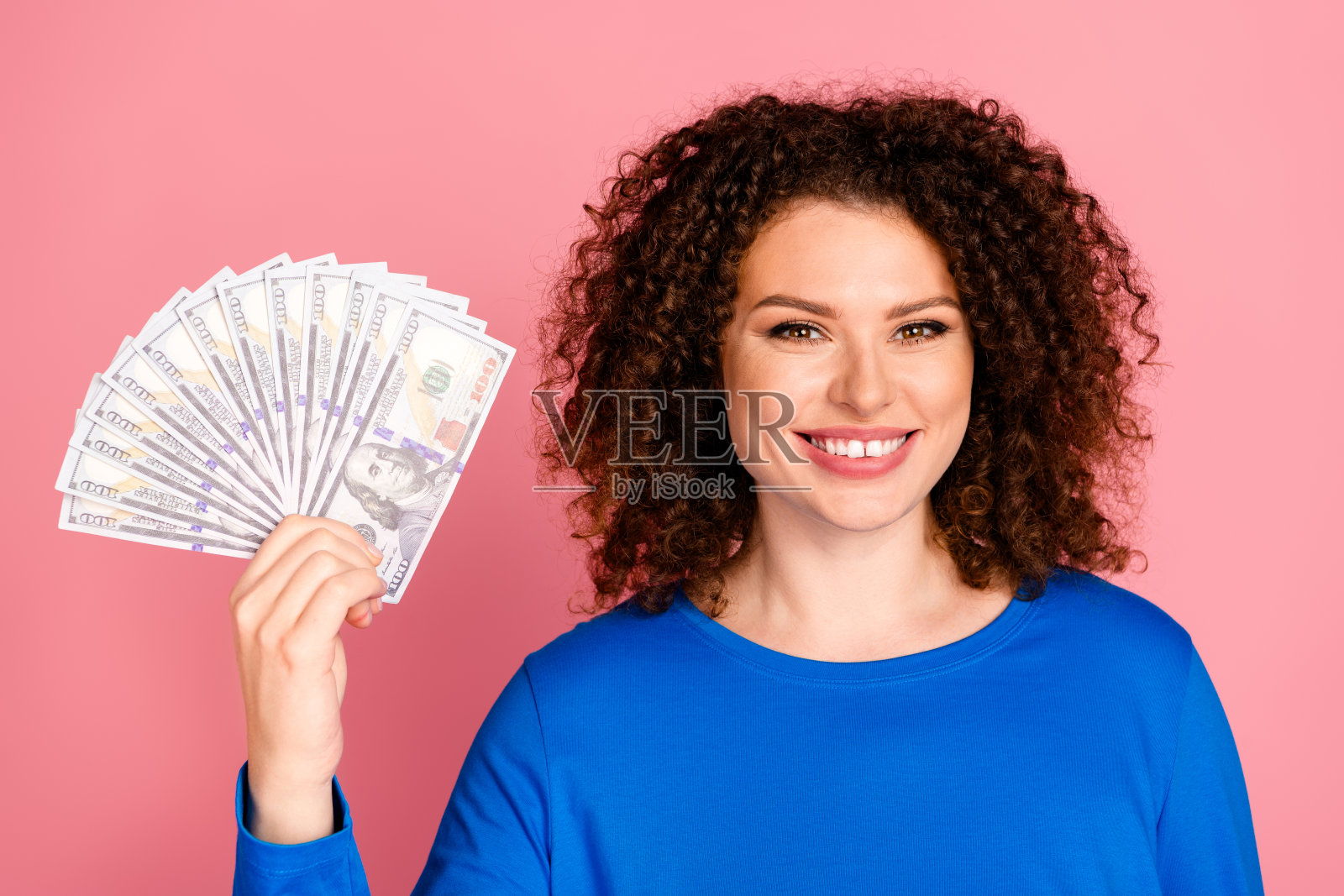 微笑的女性穿着休闲蓝色上衣，手持钞票，背景为粉色，展示商业成功和财务选择。照片摄影图片