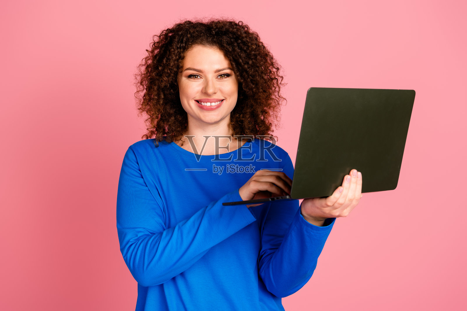 穿着休闲蓝色卫衣的快乐年轻女性在粉色背景前使用笔记本电脑，宣传在线购物和商业。照片摄影图片