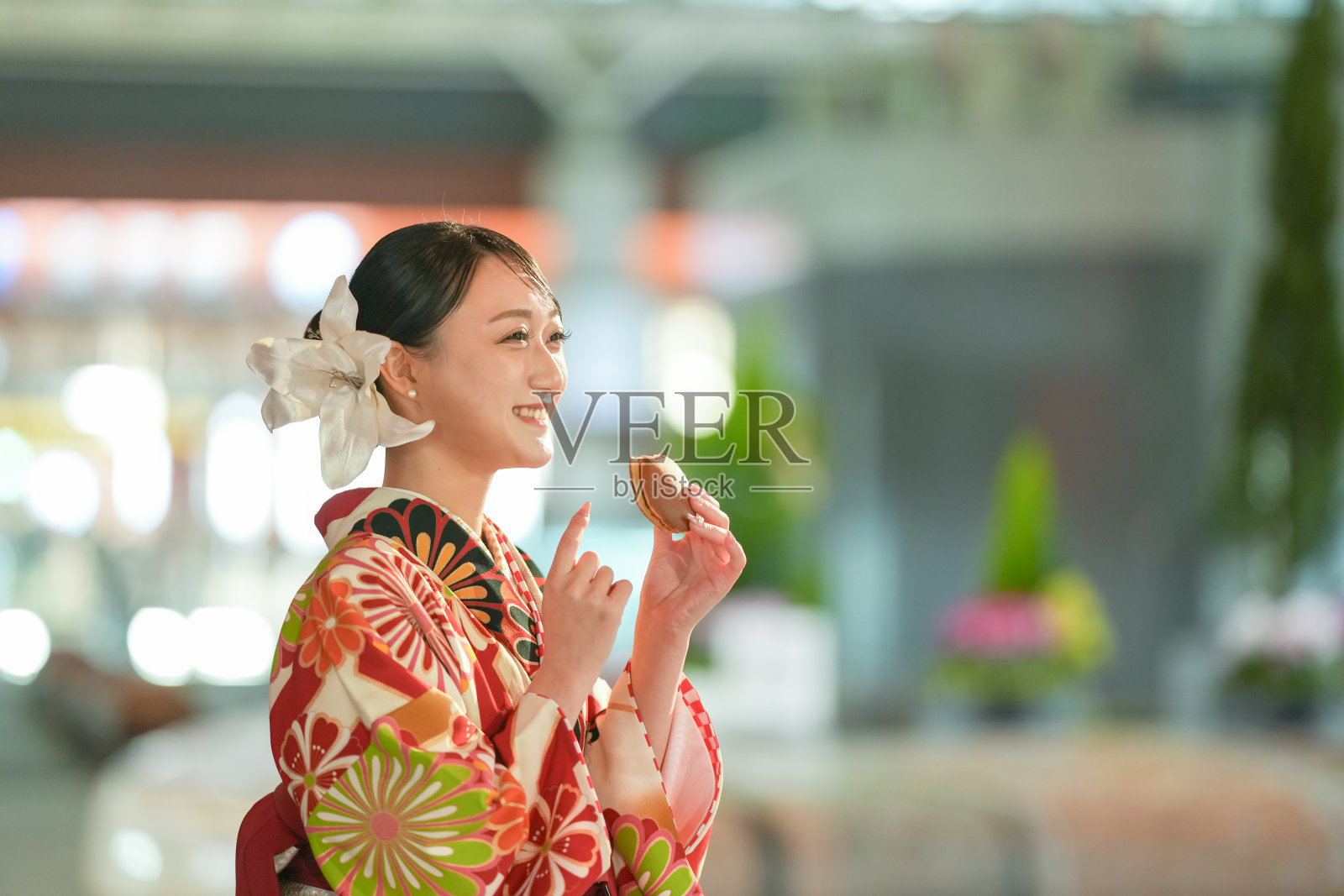 一位20多岁的日本女性，穿着红色和服，这是日本大学毕业生的标志，正在石川县金泽市的一个灯光明亮的车站吃着铜锣烧。照片摄影图片