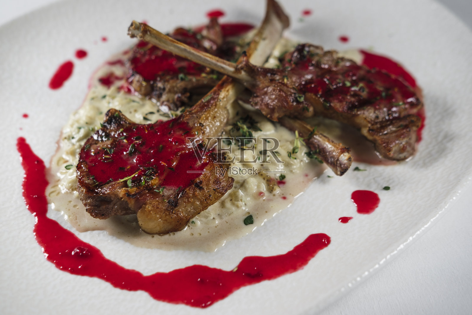 红樱桃酱烤羊排，搭配粥，撒上时令香草，盛放在白色盘子上，背景为布料。照片摄影图片
