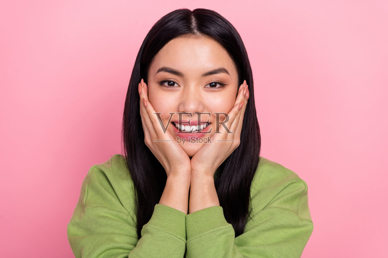 乐观的年轻女士双手托脸，穿着绿色卫衣，背景为粉色。照片摄影图片