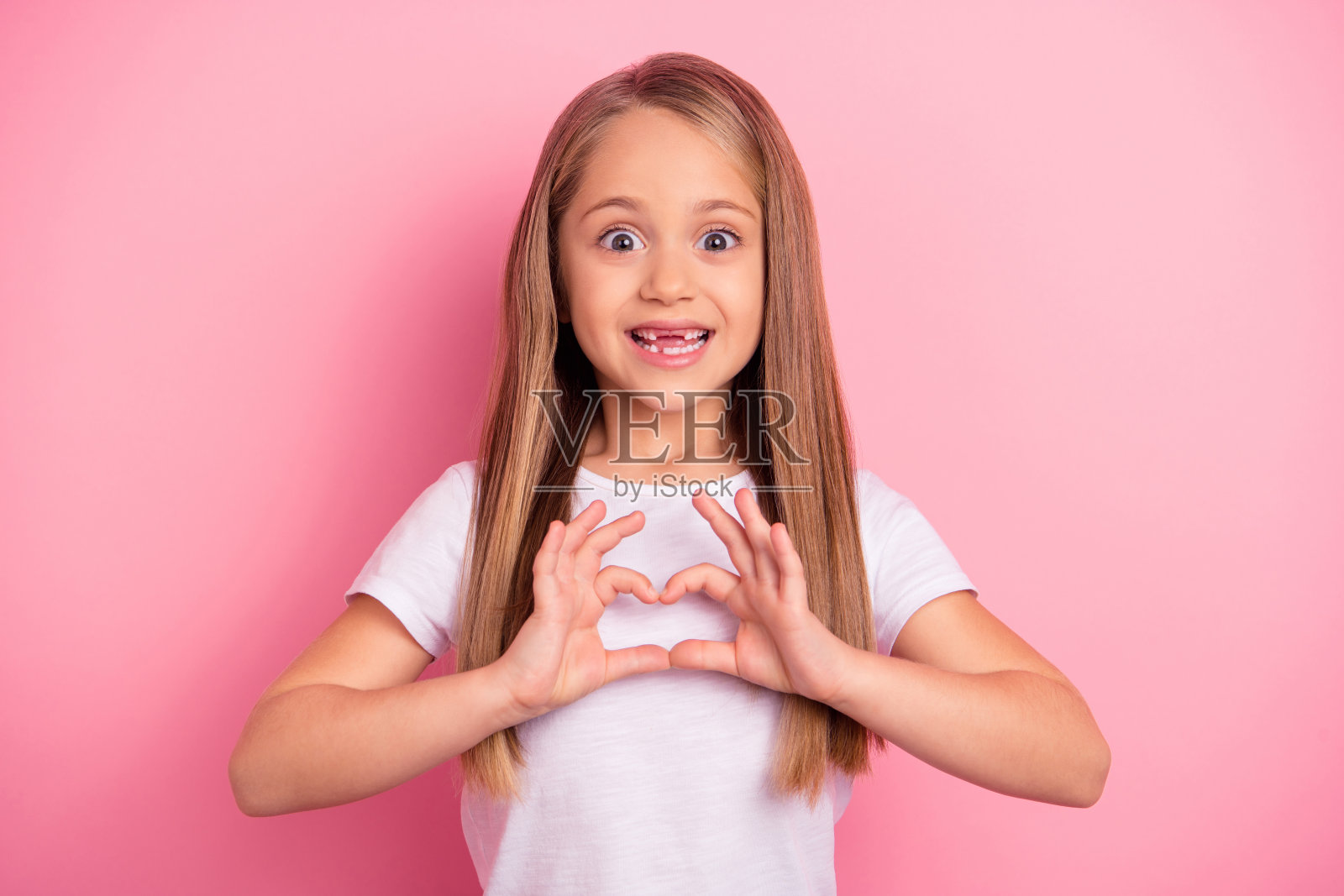 可爱女孩露齿而笑，双手比心形手势，背景为粉色。照片摄影图片
