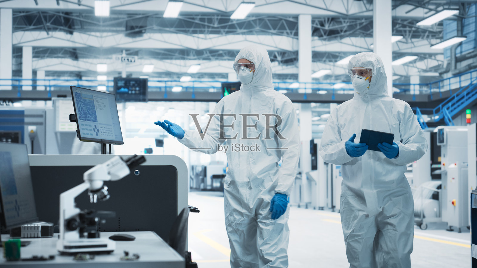 两位穿着无菌服的科学家在电子工厂研究中心走动。一名技术员手持平板电脑，工程师则走向焊接喷墨打印机，开始电路板的生产过程。照片摄影图片