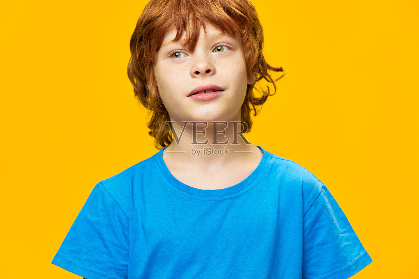 男孩的特写肖像，穿着蓝色T恤，背景为黄色，红色头发。照片摄影图片