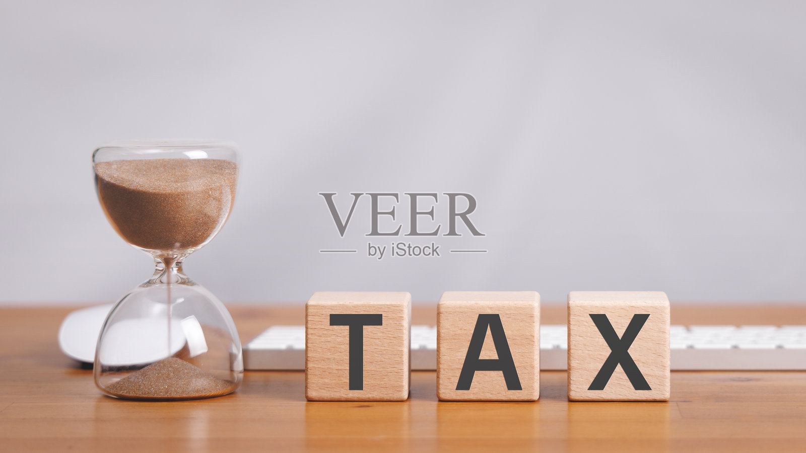 木制立方体块上有税务字样，放在木桌上，旁边有沙漏、电脑鼠标和键盘，背景为白色。照片摄影图片