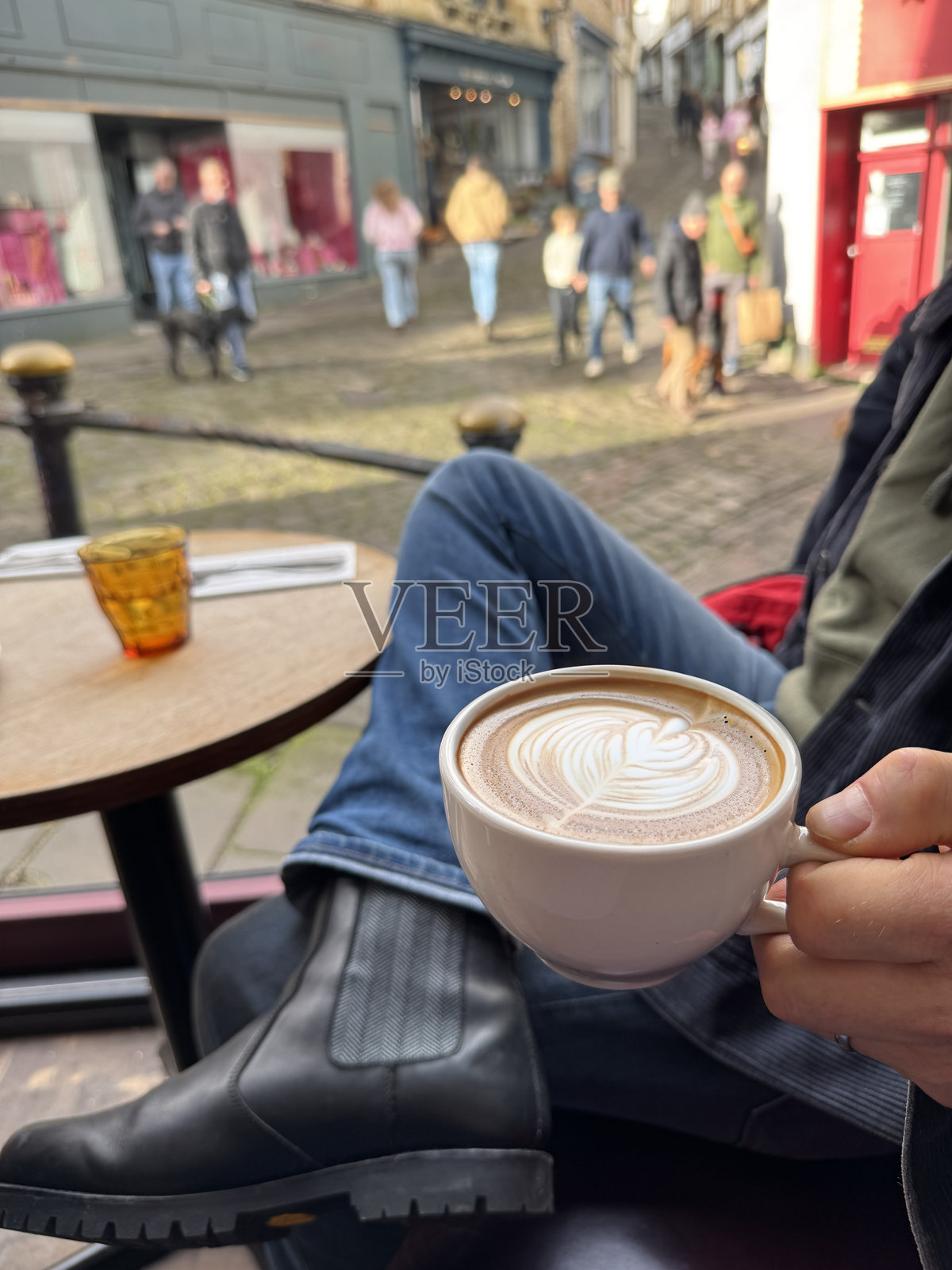 一位无法辨认的男子穿着灯芯绒夹克、蓝色牛仔裤和黑色切尔西靴，腿交叉坐在户外人行道咖啡馆的桌子旁，手中拿着一杯带有白色泡沫拉花叶子图案的咖啡，焦点在前景。照片摄影图片