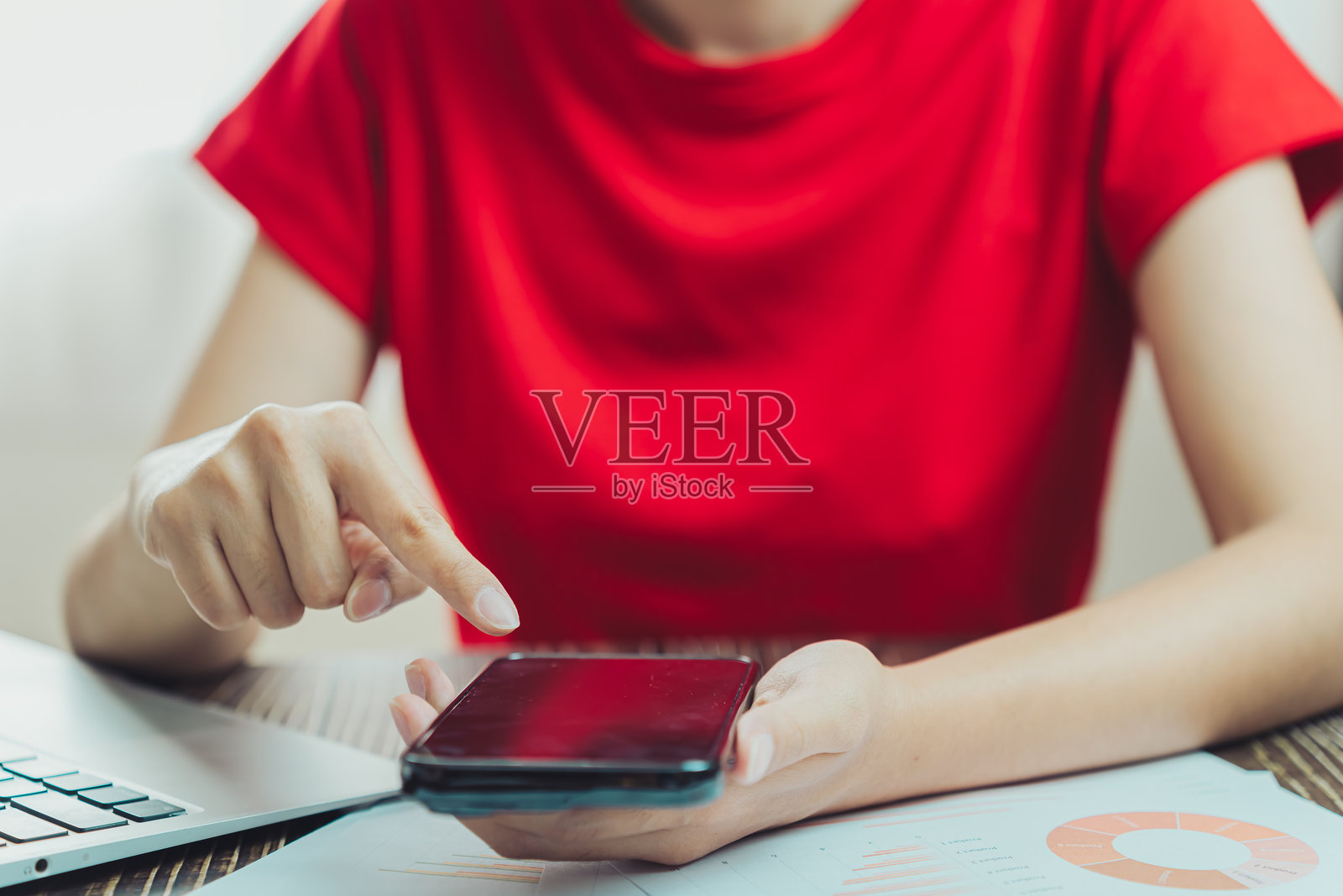 穿着红色衬衫的人坐在桌子前使用智能手机，桌上有笔记本电脑和文件。场景传达出一种高效和专注的感觉。照片摄影图片