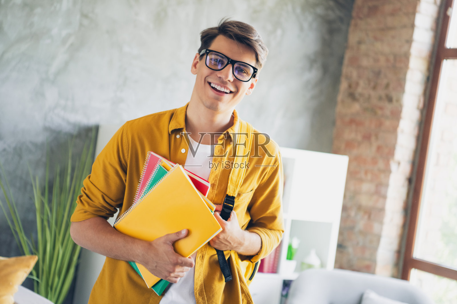 年轻男子在明亮时尚的公寓内微笑着，手里拿着五颜六色的笔记本，享受轻松的一天照片摄影图片