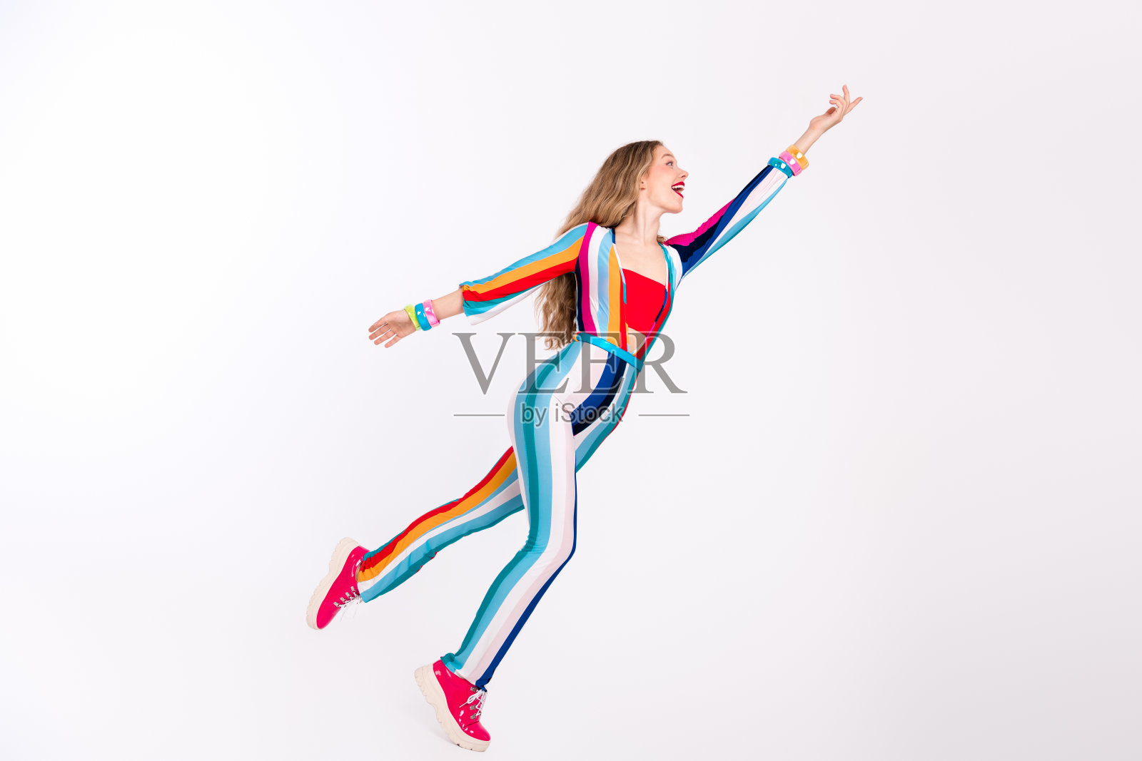 年轻女性穿着五彩条纹的连体裤，长发金黄，愉快地在白色背景前摆姿势，展现出充满活力的迪斯科氛围。照片摄影图片