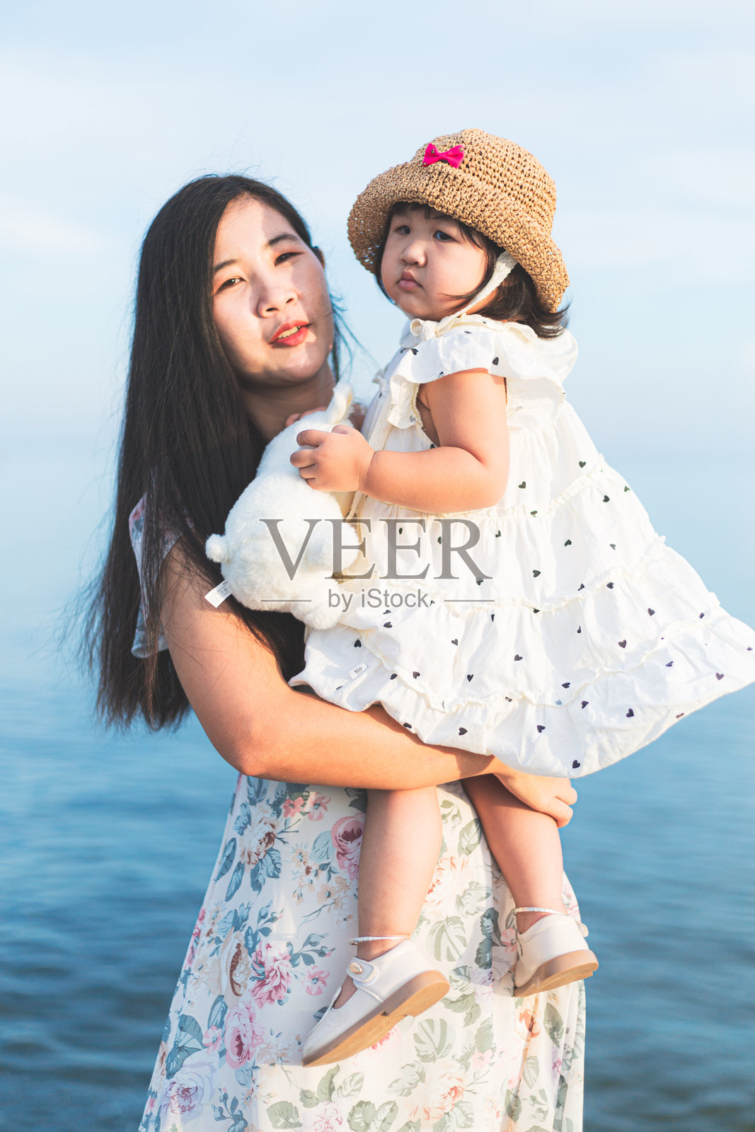 亚洲母亲在夏季日落时分，抱着并亲吻她的小女儿，走在热带海滩上。照片摄影图片