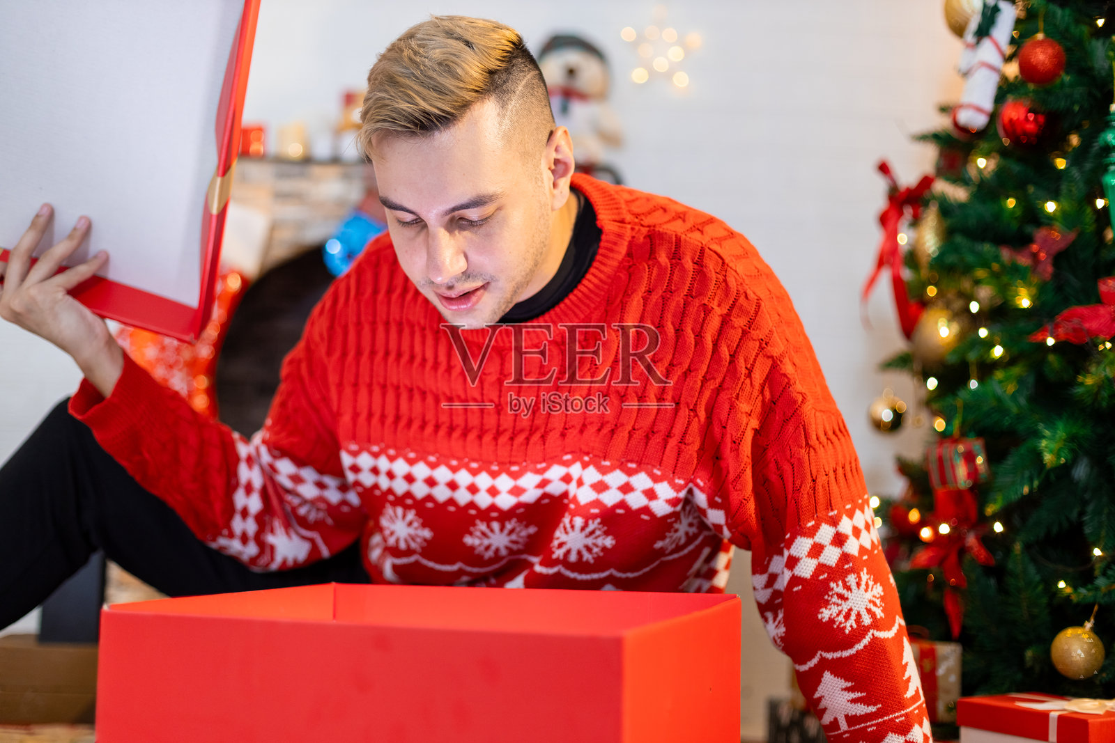 圣诞节概念。英俊的男人坐着，惊讶地打开礼物盒，背景是圣诞树。节日气氛派对。照片摄影图片