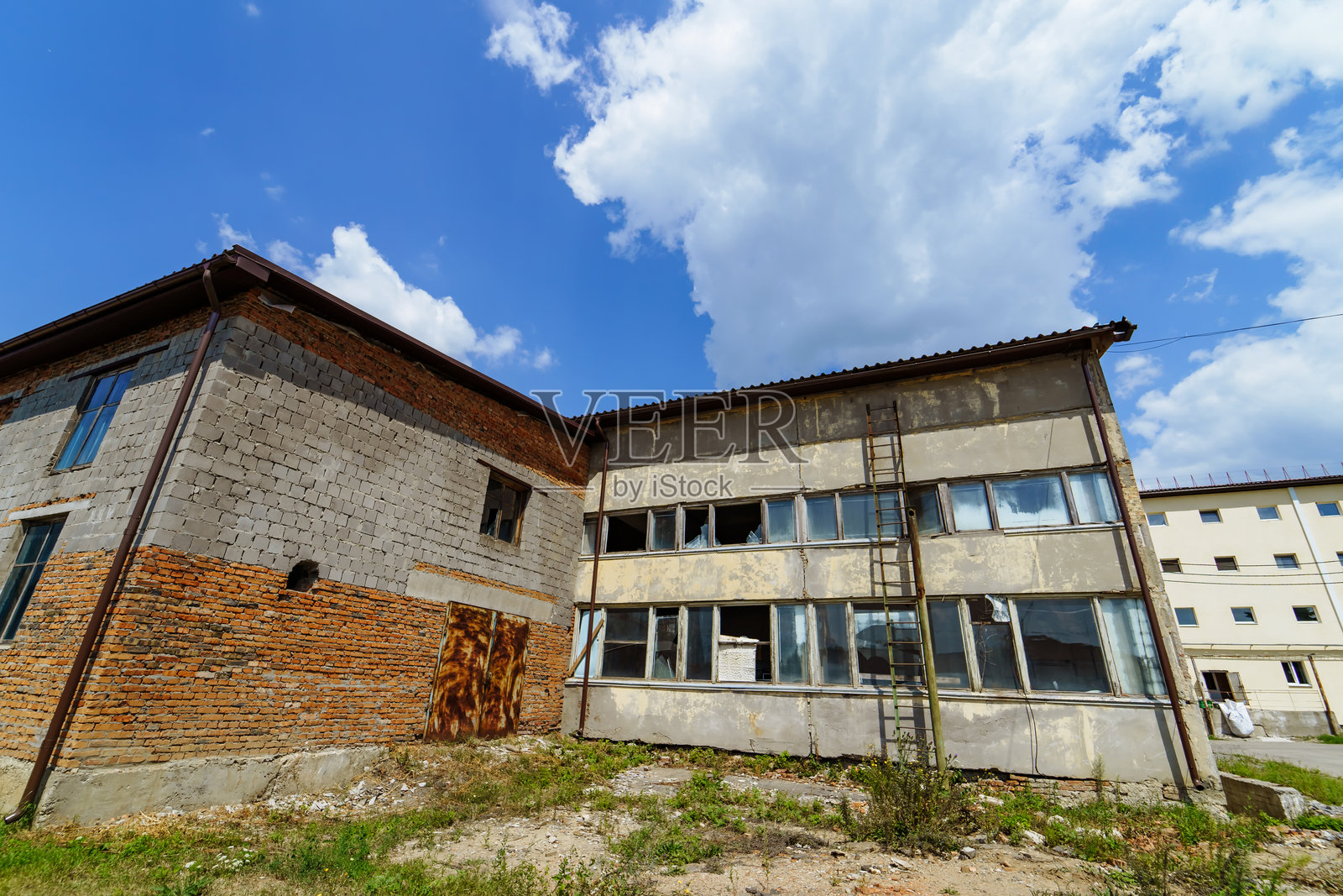在明亮的蓝天下，一座废弃的建筑物油漆剥落。老旧破败的建筑显示出剥落的油漆和一架生锈的梯子，背景是蓬松的云朵和蓝天。照片摄影图片
