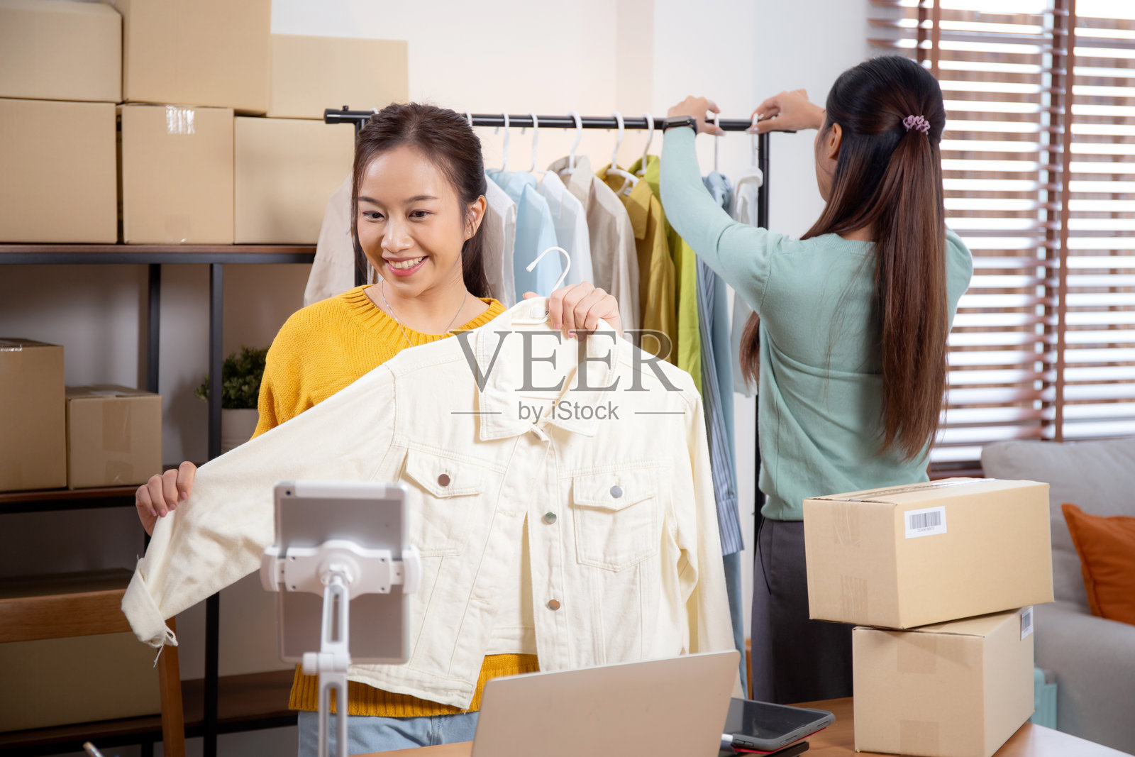 两位年轻的亚洲女性在家中通过平板电脑和笔记本电脑展示服装，经营在线服装店，作为企业家进行直播销售，展示在线销售服装的过程。照片摄影图片