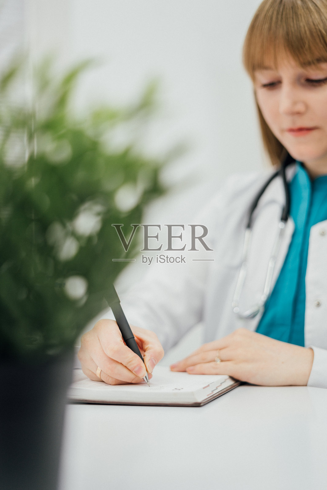 女性医生在白色桌子上用夹板写字的特写。医生穿着白大褂，脖子上挂着听诊器，突显出专业的医疗环境。照片摄影图片