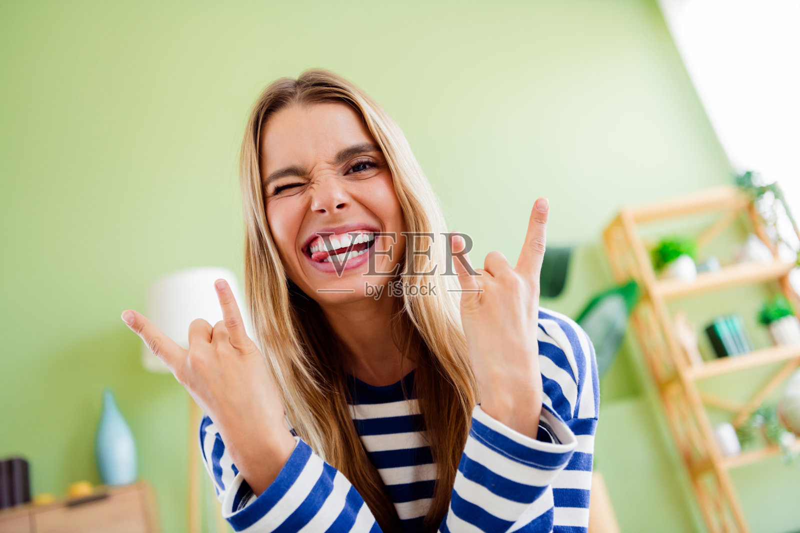 可爱的金发小女孩在宽敞的室内房间里穿着条纹衣服，做出摇滚手势。照片摄影图片