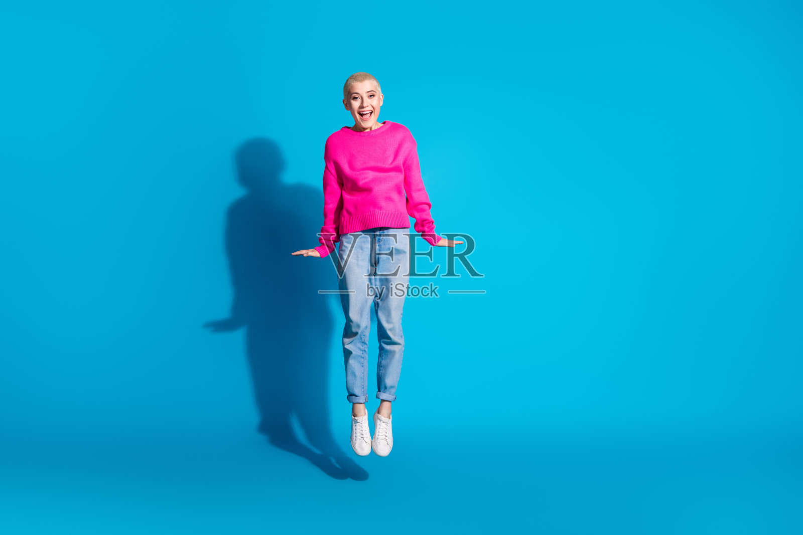 年轻女性在明亮的蓝色背景下，穿着鲜艳的粉色毛衣欢快地跳跃，展现出风格和情感。照片摄影图片
