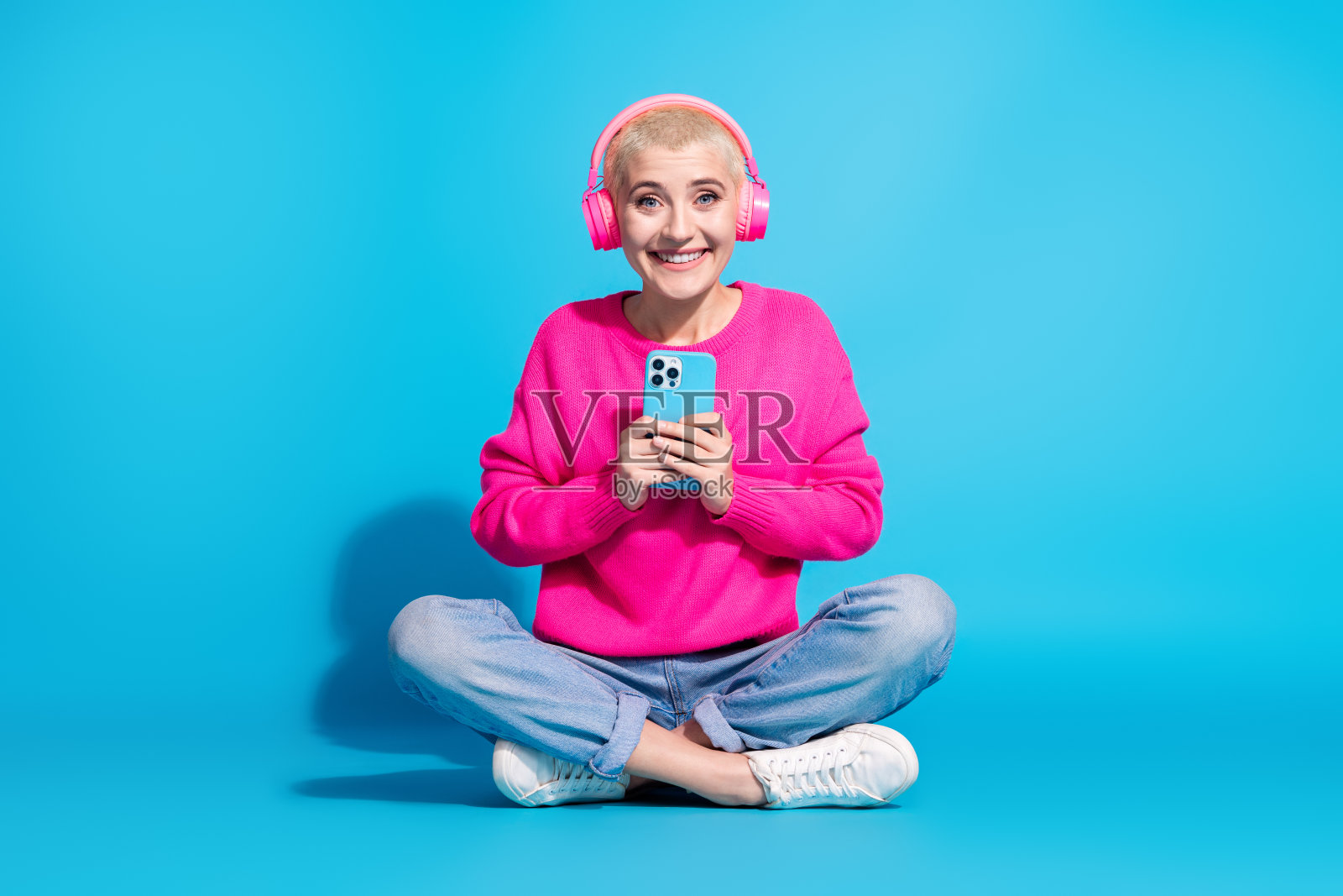 年轻女性穿着粉色毛衣，戴着耳机，盘腿坐在蓝色背景前，手持手机，面带微笑。照片摄影图片