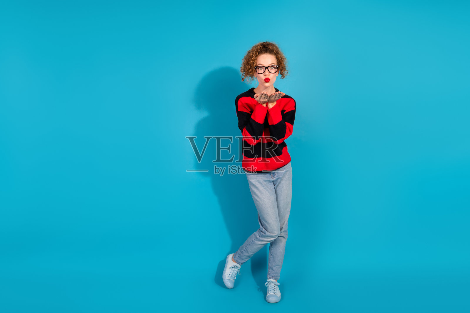 年轻时尚的卷发女性穿着条纹毛衣，摆姿势于鲜艳的蓝色摄影棚背景前。照片摄影图片