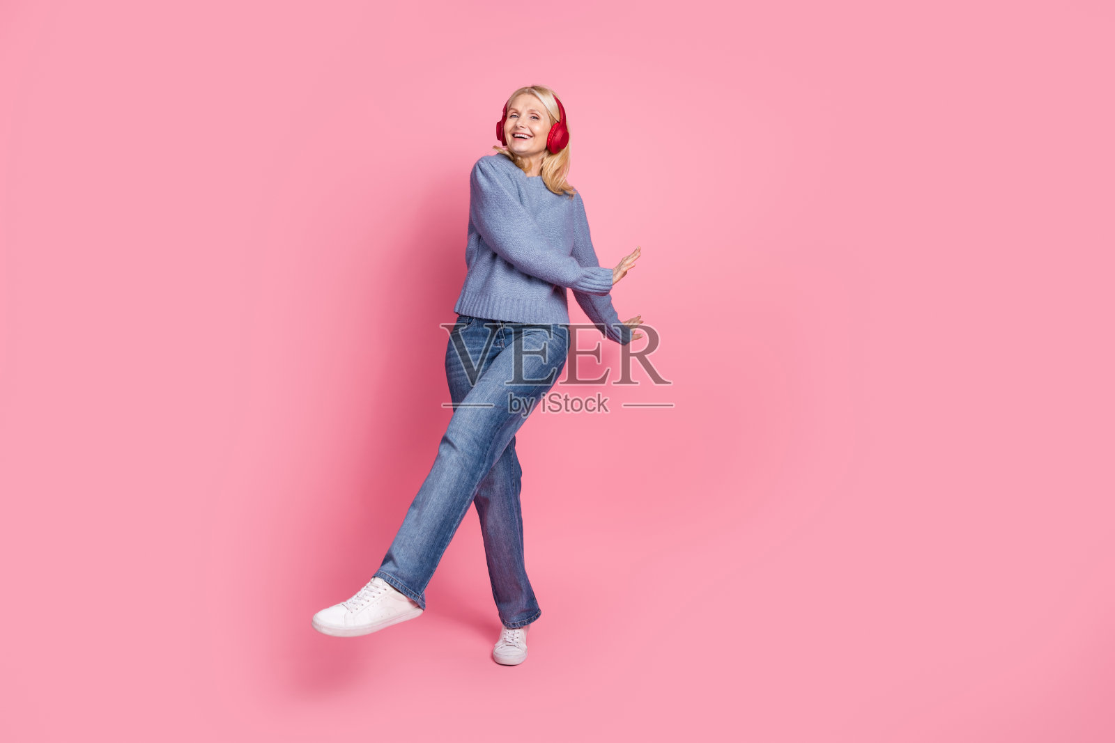 全身照，迷人的老年女性穿着时尚的蓝色针织服装，戴着耳机，快乐地跳舞，背景为粉色。照片摄影图片