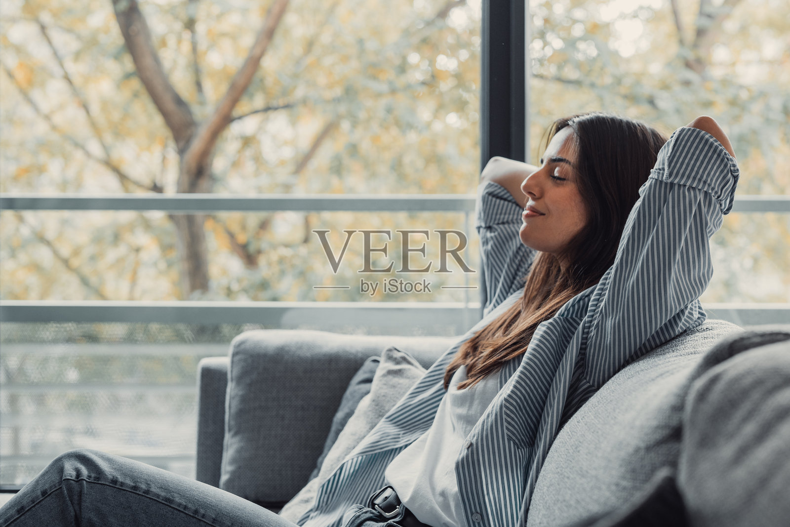 放松宁静的年轻女性感到疲惫，双手放在头后，舒适地躺在沙发上，快乐而平静的女士在现代客厅中享受健康，呼吸新鲜空气。照片摄影图片