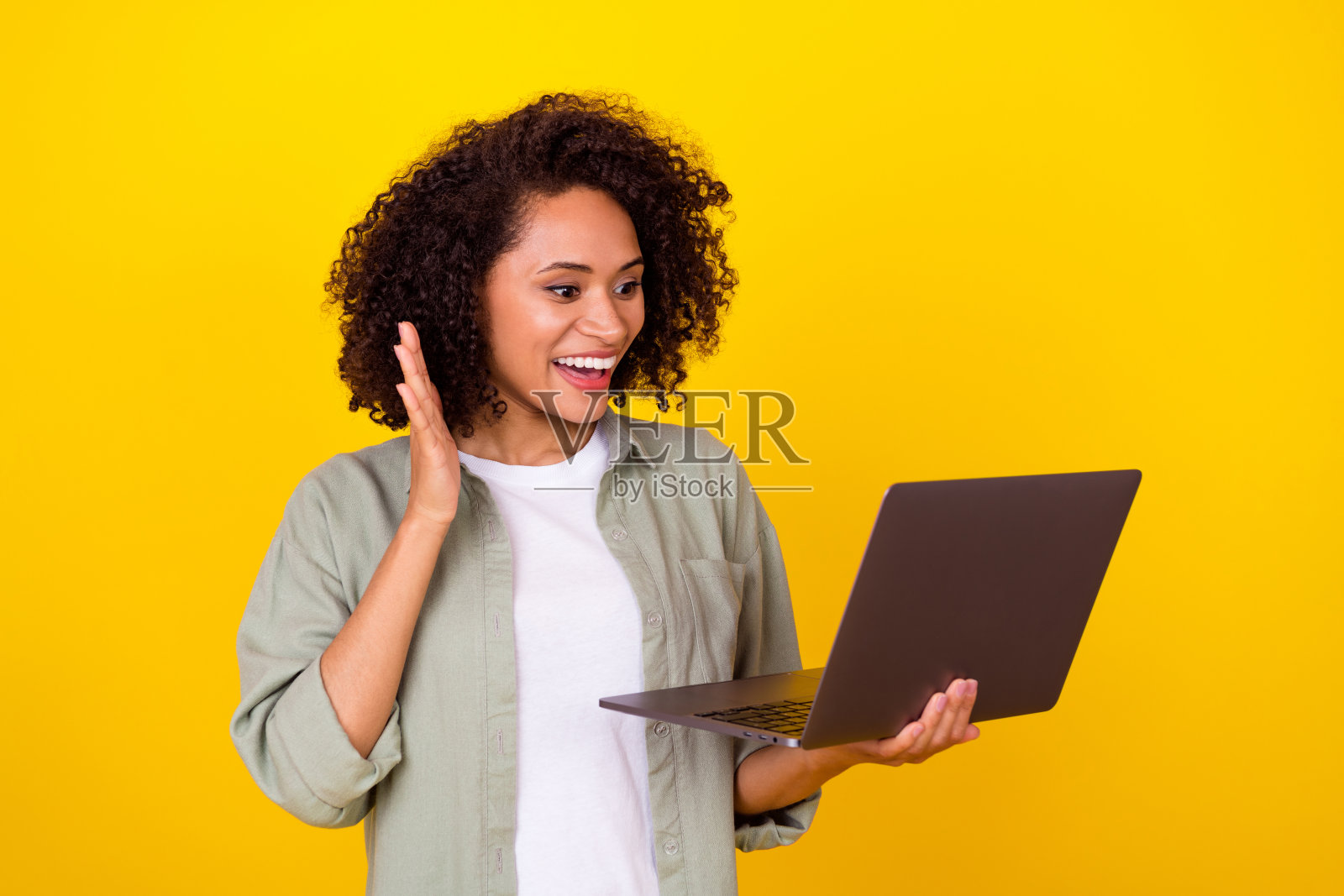 一位印象深刻的千禧世代棕发女士穿着灰色衬衫，坐在黄色背景下与笔记本电脑交谈。照片摄影图片