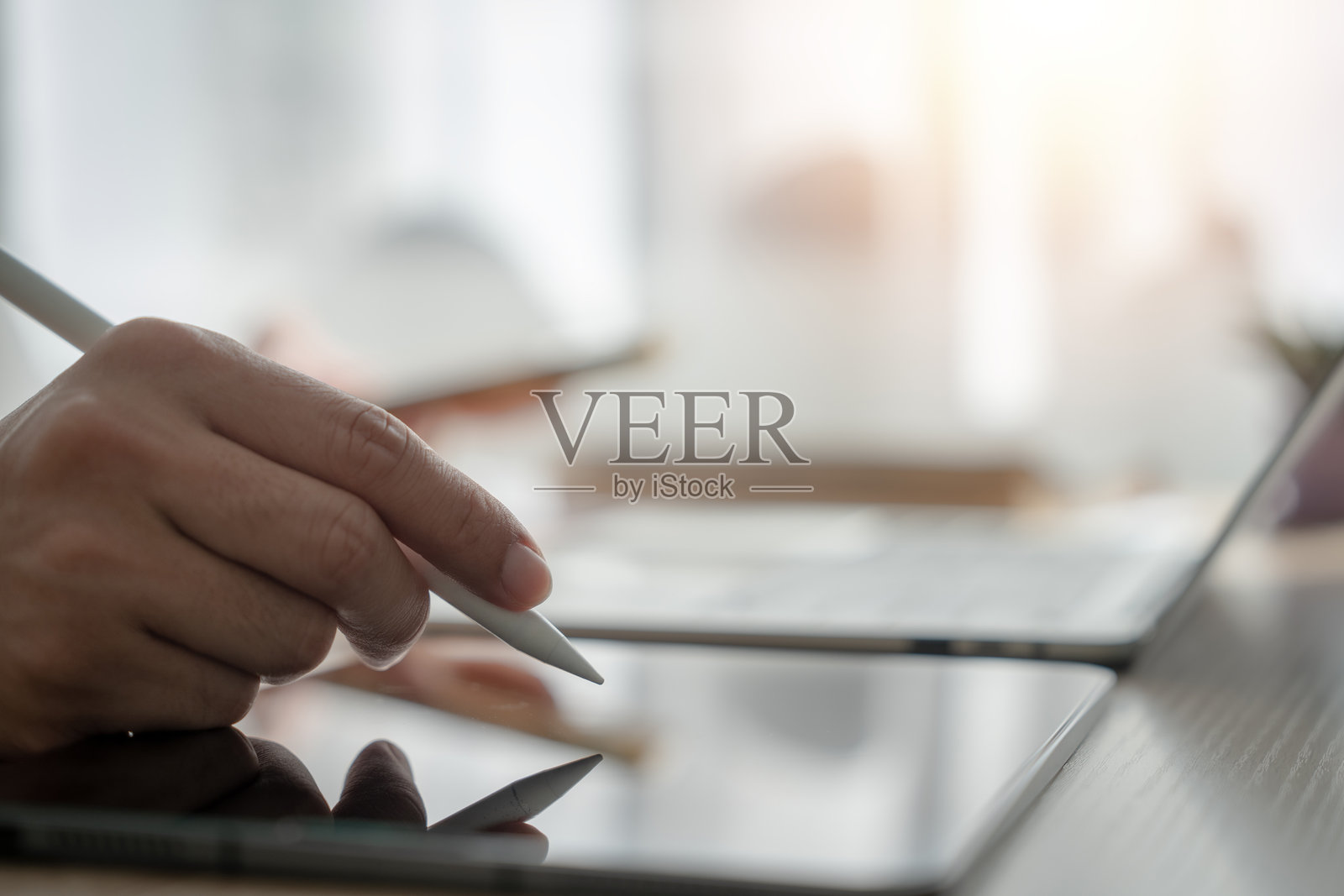 一个人正在使用平板电脑和手写笔进行书写。平板电脑放在桌子上，桌子上还有其他物品，如笔记本电脑和手机。照片摄影图片