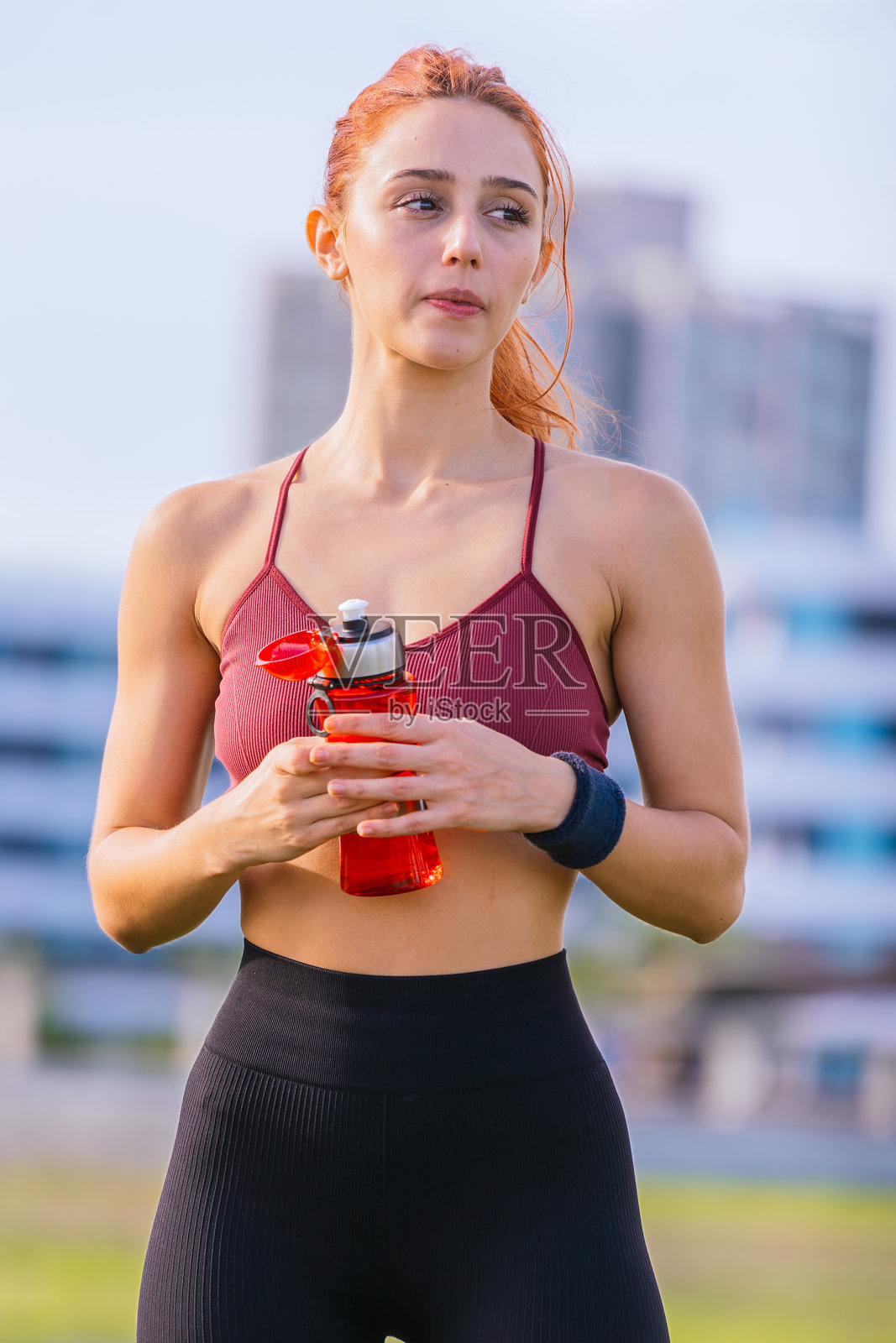 在阳光明媚的城市公园里，一位运动女性手持红色水瓶。照片摄影图片