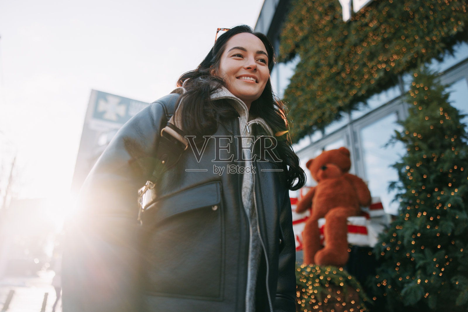 年轻微笑的女性站在城市街道上，旁边是装饰着闪亮灯串的圣诞树。照片摄影图片