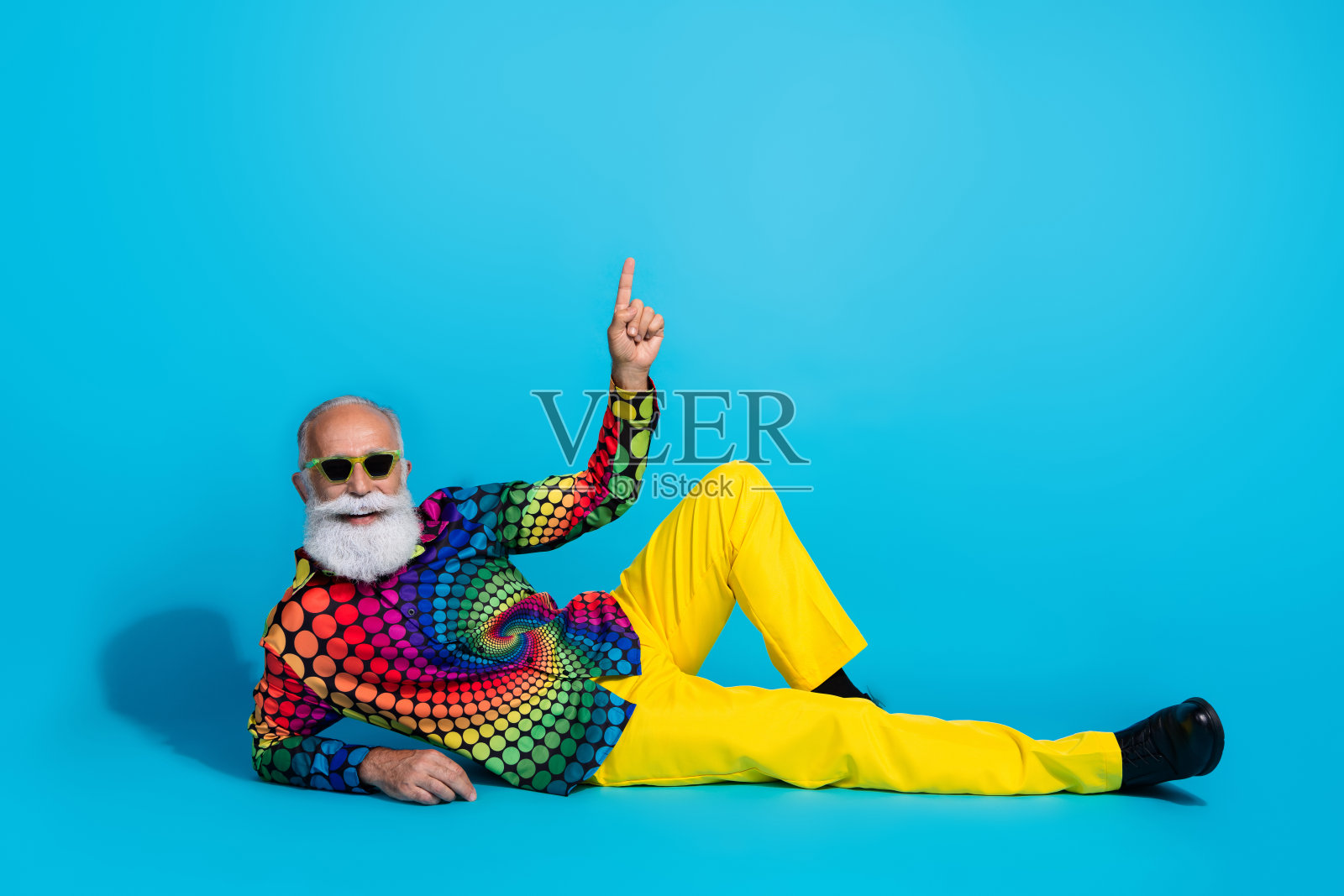 全身照：一位幽默时尚的老年男子坐在地板上，手指指向空白空间，背景为蓝色。照片摄影图片