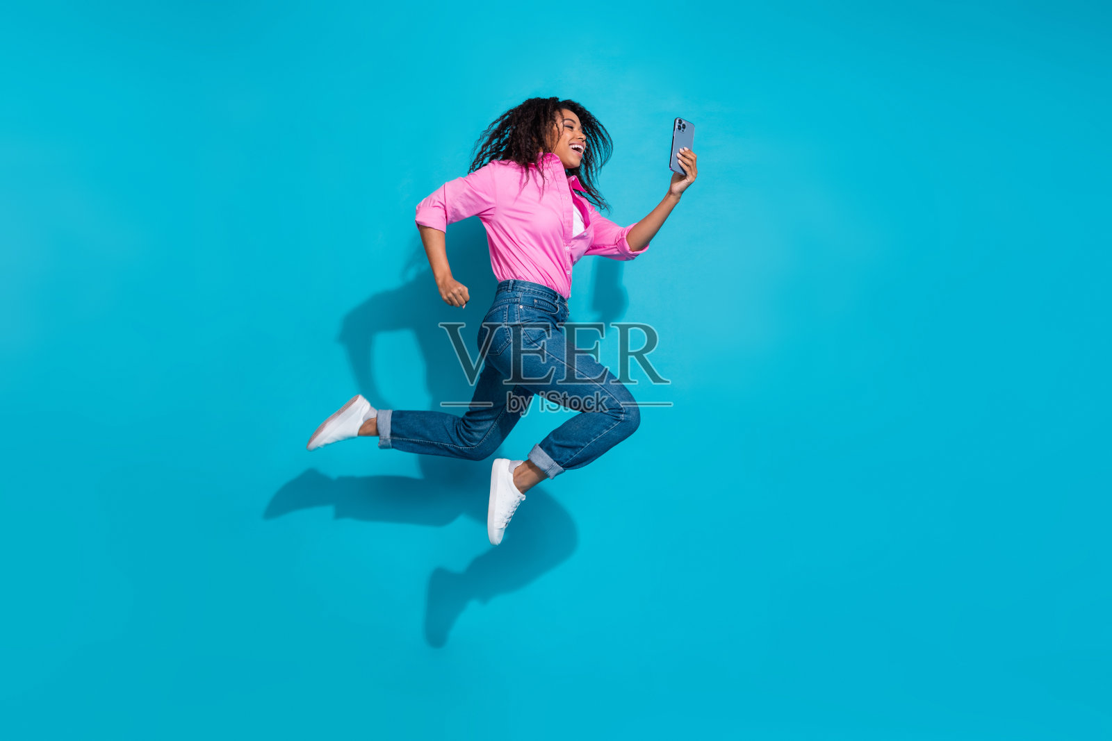 穿着粉色衬衫和牛仔裤的快乐年轻女性在鲜艳的蓝色背景前跳跃，捕捉到幸福与时尚的自拍时刻。照片摄影图片