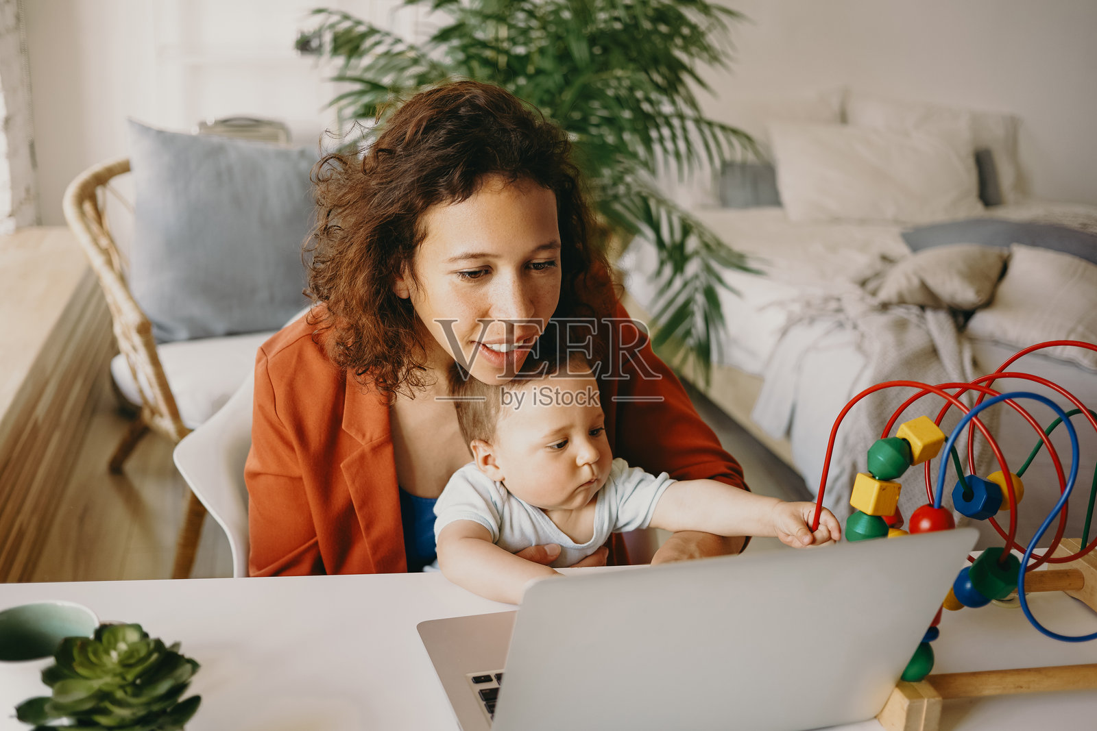 快乐成功的年轻女商人兼顾事业与母亲角色，在家中使用普通笔记本电脑远程工作，膝上坐着她的幼儿儿子，正在玩玩具。照片摄影图片